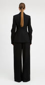 A model viewed from the back wearing a black tailored suit with a structured blazer and wide-leg trousers, standing against a plain light gray studio background.