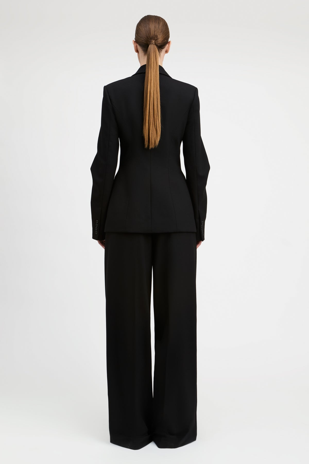 A model viewed from the back wearing a black tailored suit with a structured blazer and wide-leg trousers, standing against a plain light gray studio background.