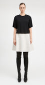 A model stands against a light gray studio background wearing a black short-sleeve cropped top, a textured off-white flared skirt, black thigh-high boots, and black pointed shoes.