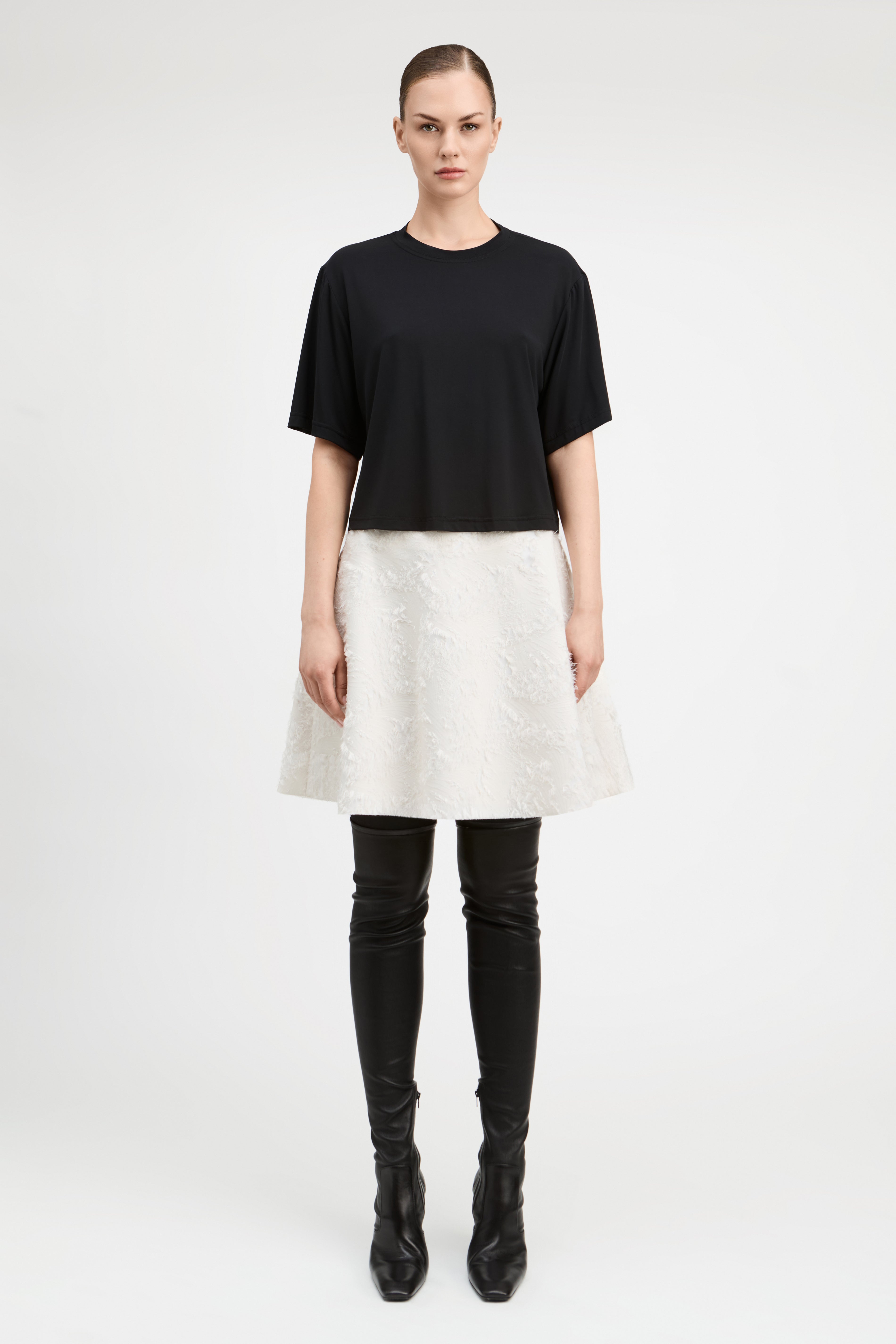 A model stands against a light gray studio background wearing a black short-sleeve cropped top, a textured off-white flared skirt, black thigh-high boots, and black pointed shoes.