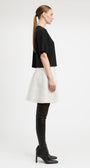 A model stands against a light gray studio background wearing a black short-sleeve cropped top, a textured off-white flared skirt, black thigh-high boots, and black pointed shoes.