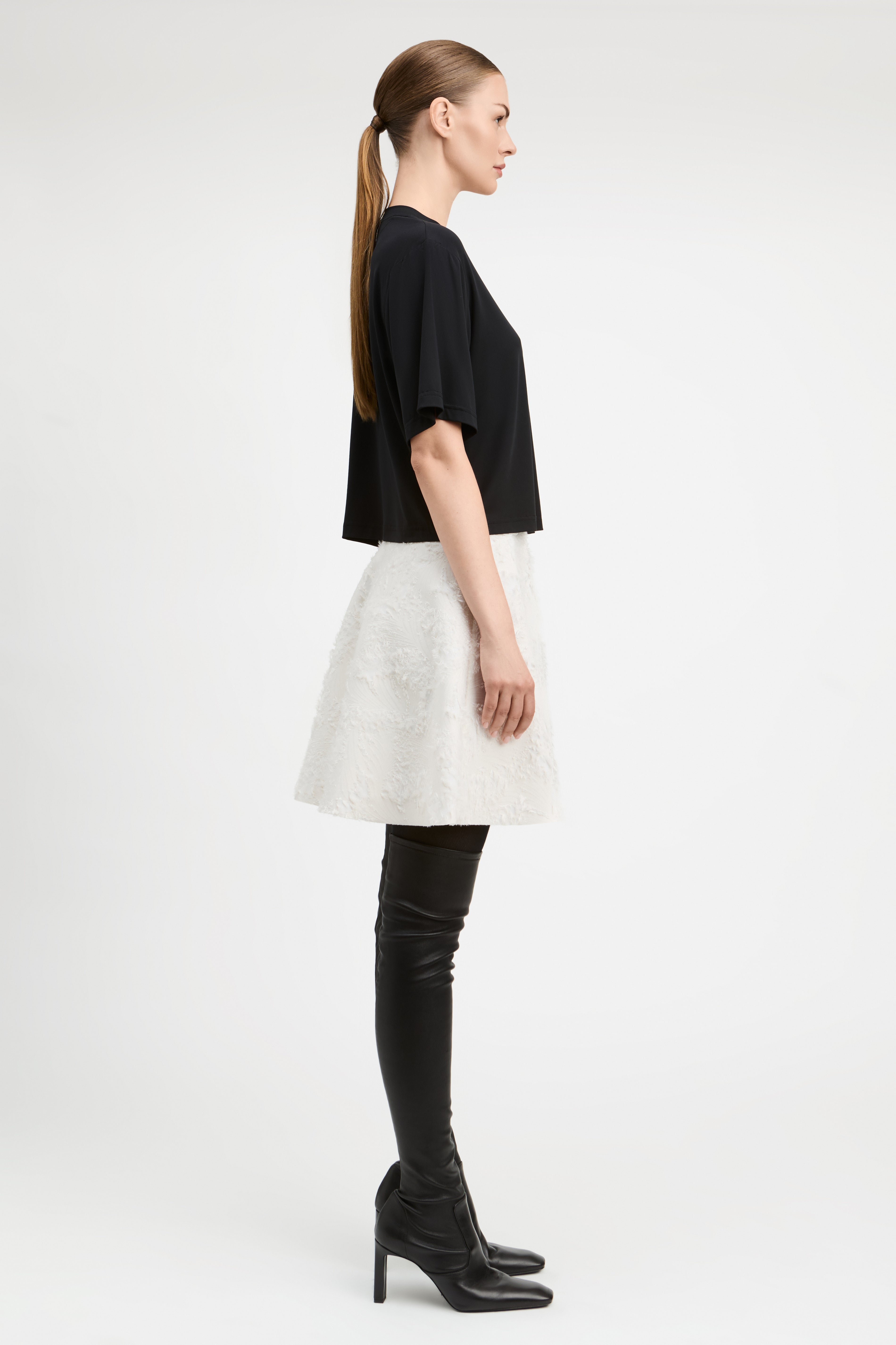 A model stands against a light gray studio background wearing a black short-sleeve cropped top, a textured off-white flared skirt, black thigh-high boots, and black pointed shoes.
