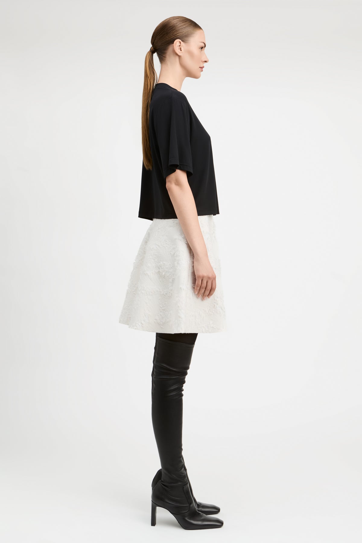 A model stands against a light gray studio background wearing a black short-sleeve cropped top, a textured off-white flared skirt, black thigh-high boots, and black pointed shoes.