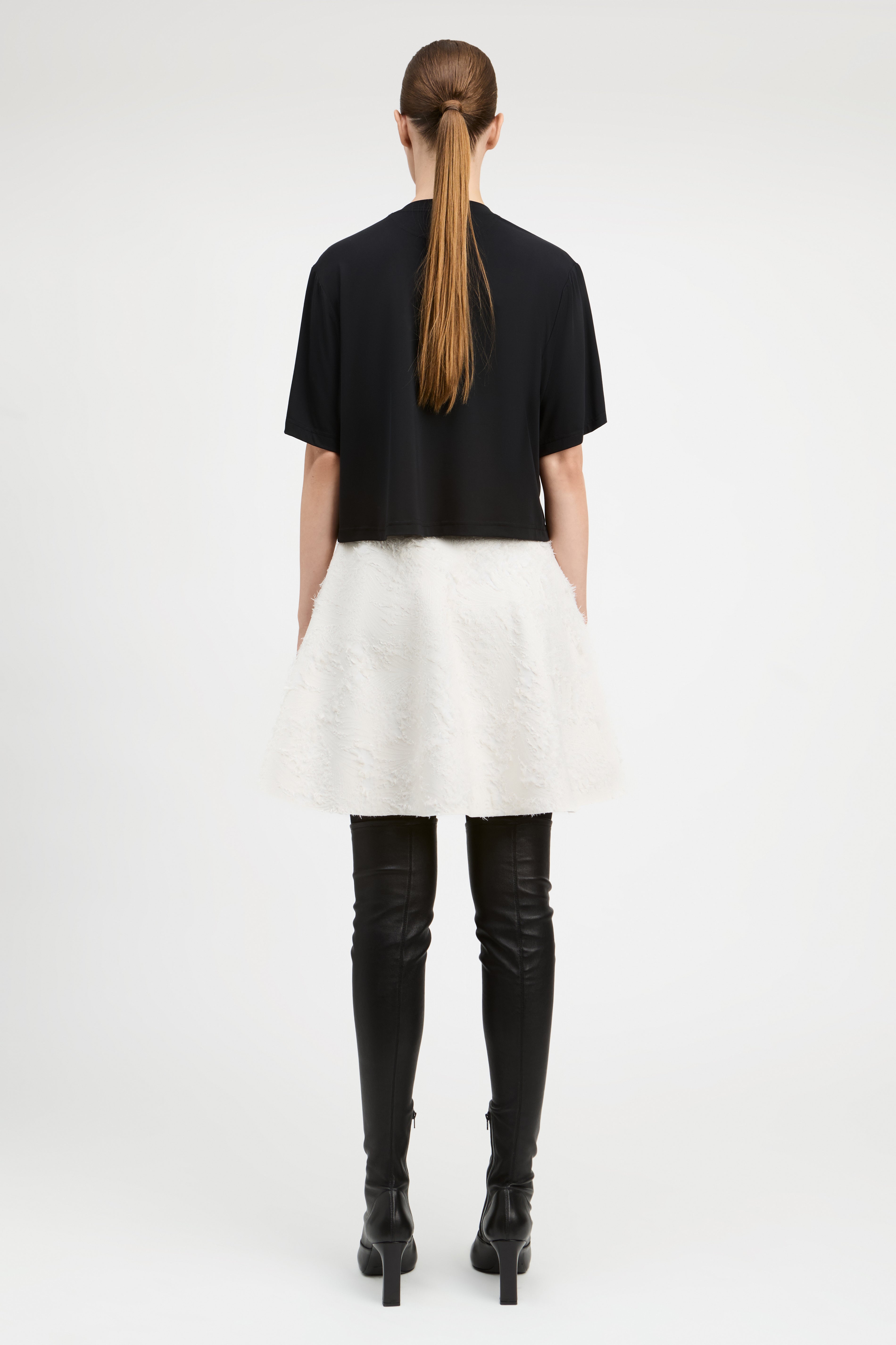 A model shown from the back wearing a black short-sleeve cropped top, a fluffy white mini skirt, black leather-look over-the-knee boots, and black high heels against a plain white studio background.