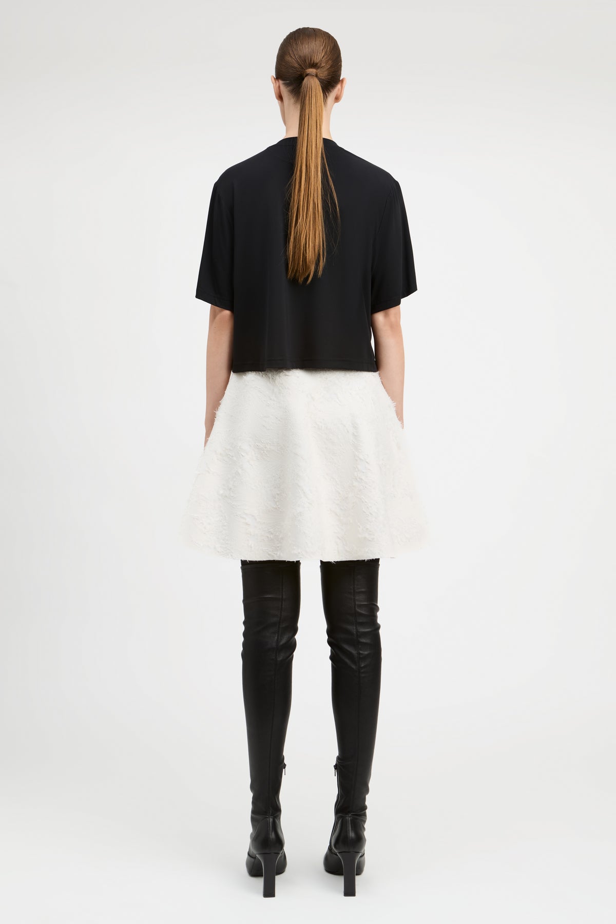 A model shown from the back wearing a black short-sleeve cropped top, a fluffy white mini skirt, black leather-look over-the-knee boots, and black high heels against a plain white studio background.