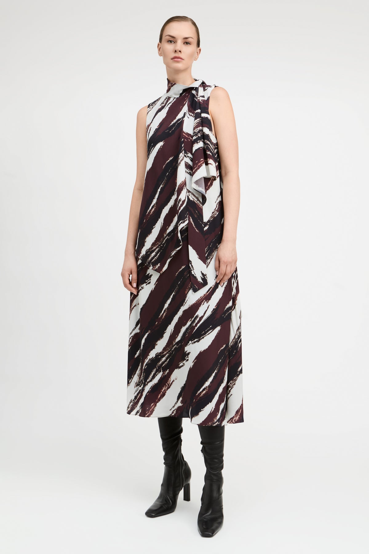 A model stands against a light gray studio background wearing a sleeveless high-neck midi dress with an abstract brushstroke pattern in dark burgundy, black, and off-white. She is also wearing black knee-high boots.