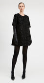 A model stands against a light gray studio background wearing a black textured mini dress with short sleeves, black leather gloves, black tights, and black pointed heels.