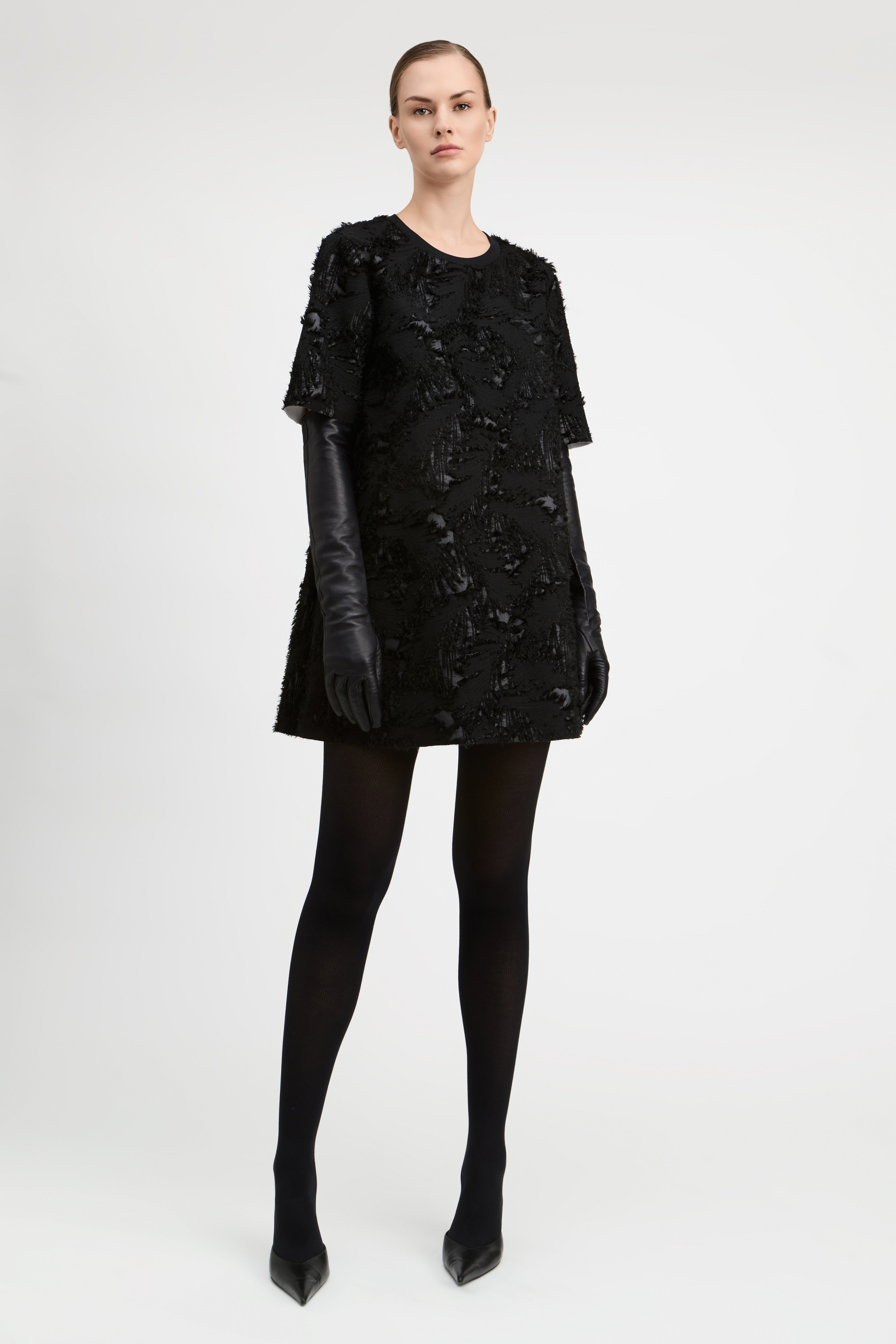 A model stands against a light gray studio background wearing a black textured mini dress with short sleeves, black leather gloves, black tights, and black pointed heels.