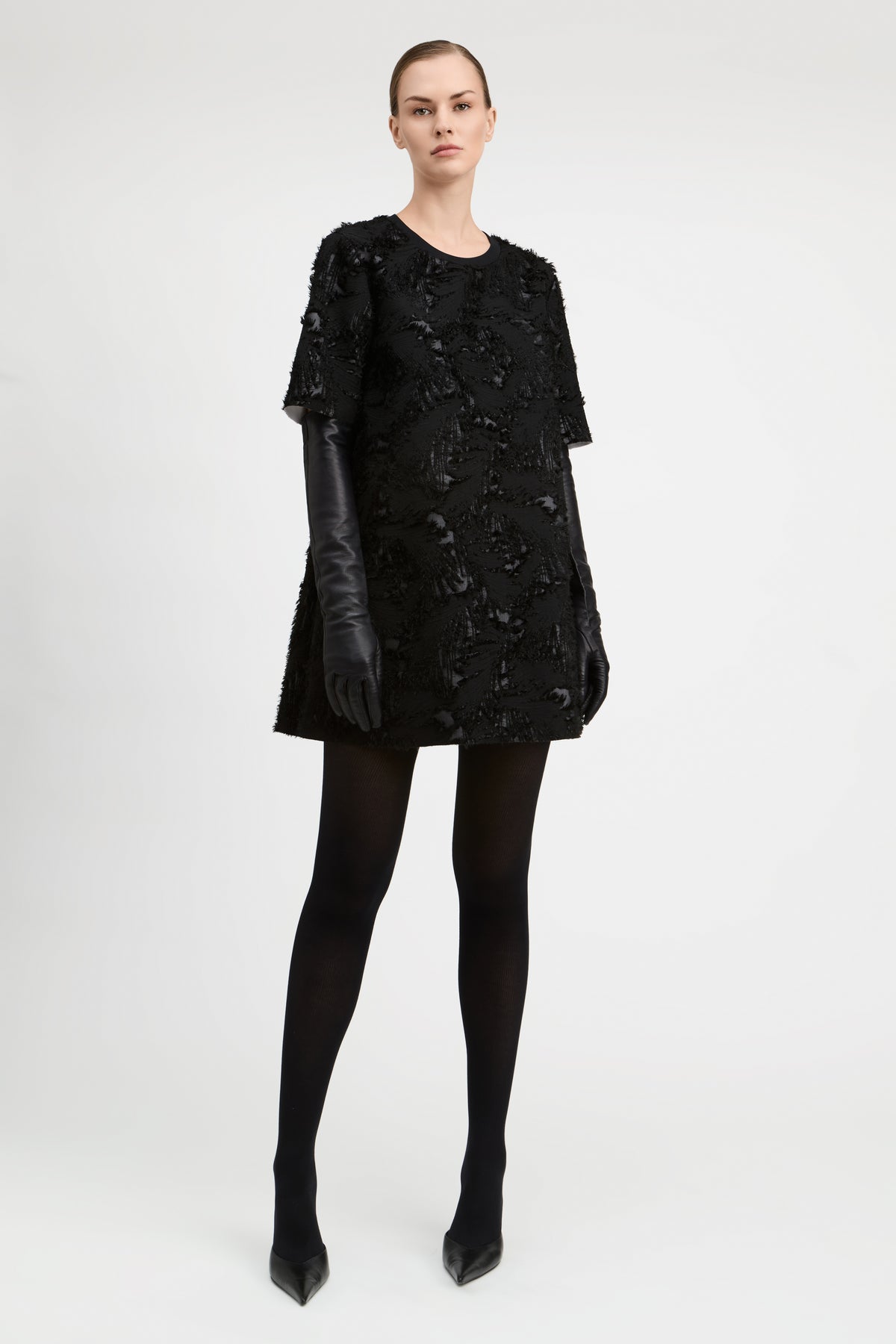 A model stands against a light gray studio background wearing a black textured mini dress with short sleeves, black leather gloves, black tights, and black pointed heels.