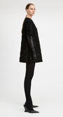 A model is shown in profile wearing a black textured, heavily embellished short coat with long black leather sleeves, paired with black tights and black pointed-toe heels, against a plain light gray studio background.