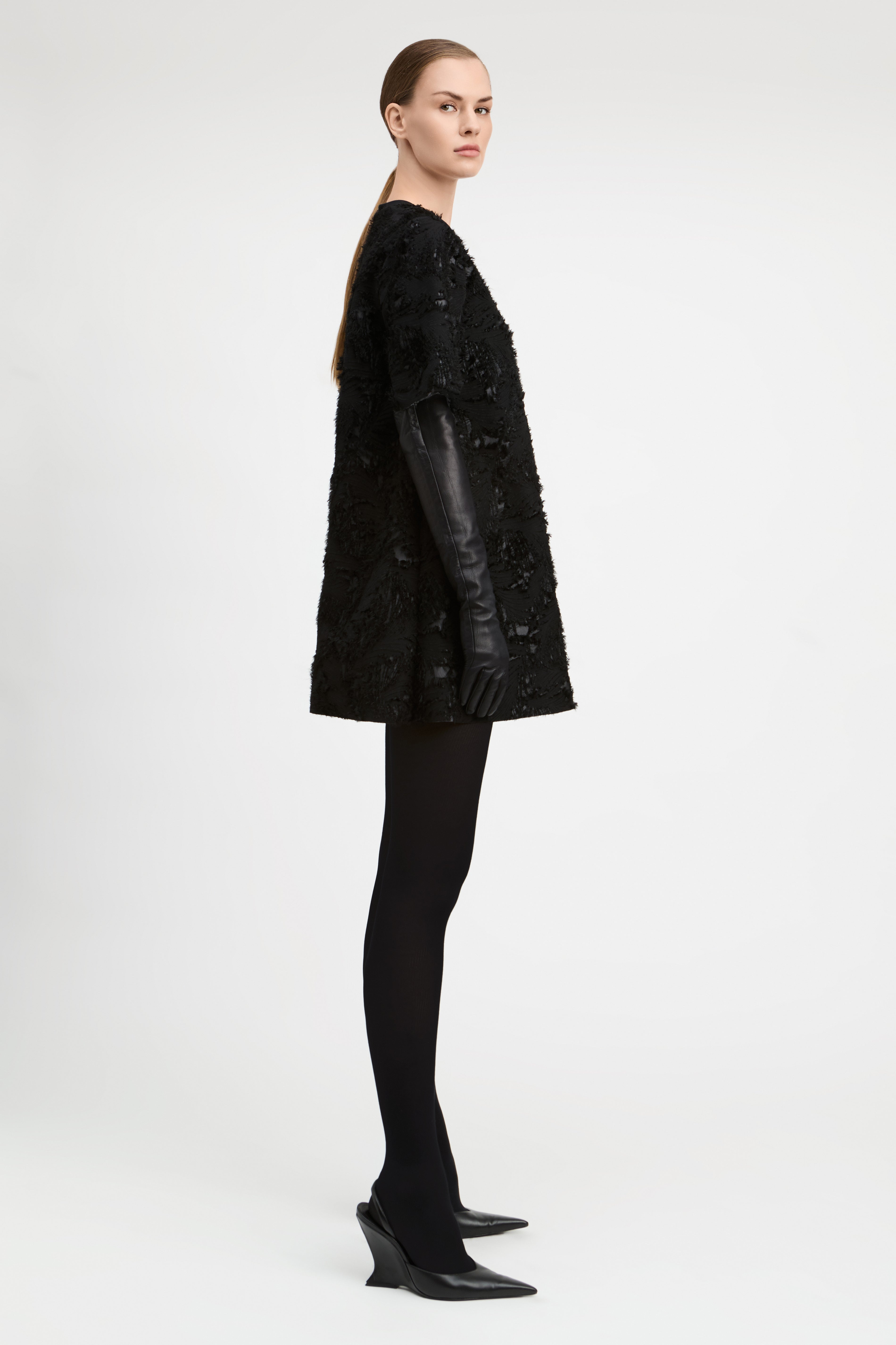 A model is shown in profile wearing a black textured, heavily embellished short coat with long black leather sleeves, paired with black tights and black pointed-toe heels, against a plain light gray studio background.