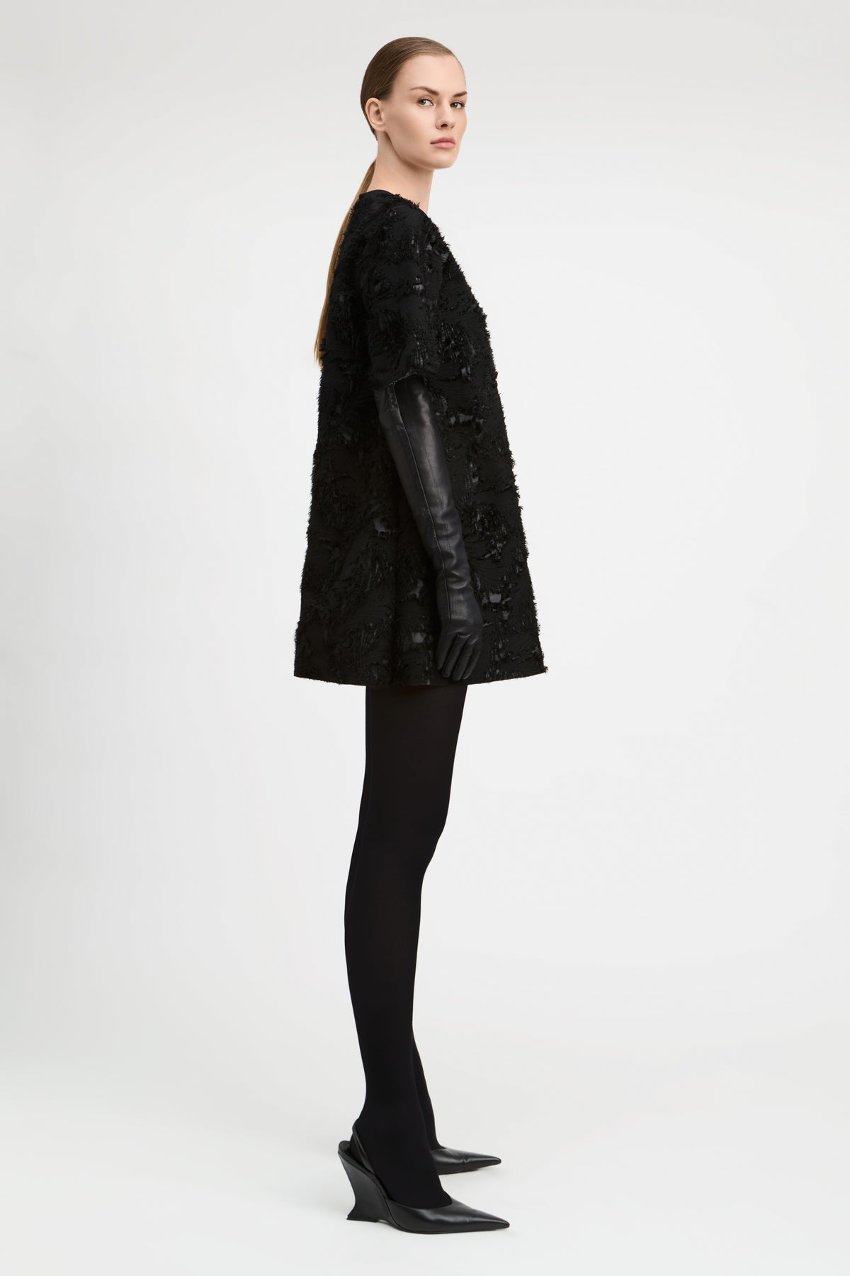A model is shown in profile wearing a black textured, heavily embellished short coat with long black leather sleeves, paired with black tights and black pointed-toe heels, against a plain light gray studio background.