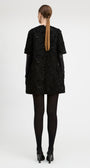 A woman viewed from the back wearing a short black textured dress with black gloves, black tights, and black heels against a plain light gray studio background.