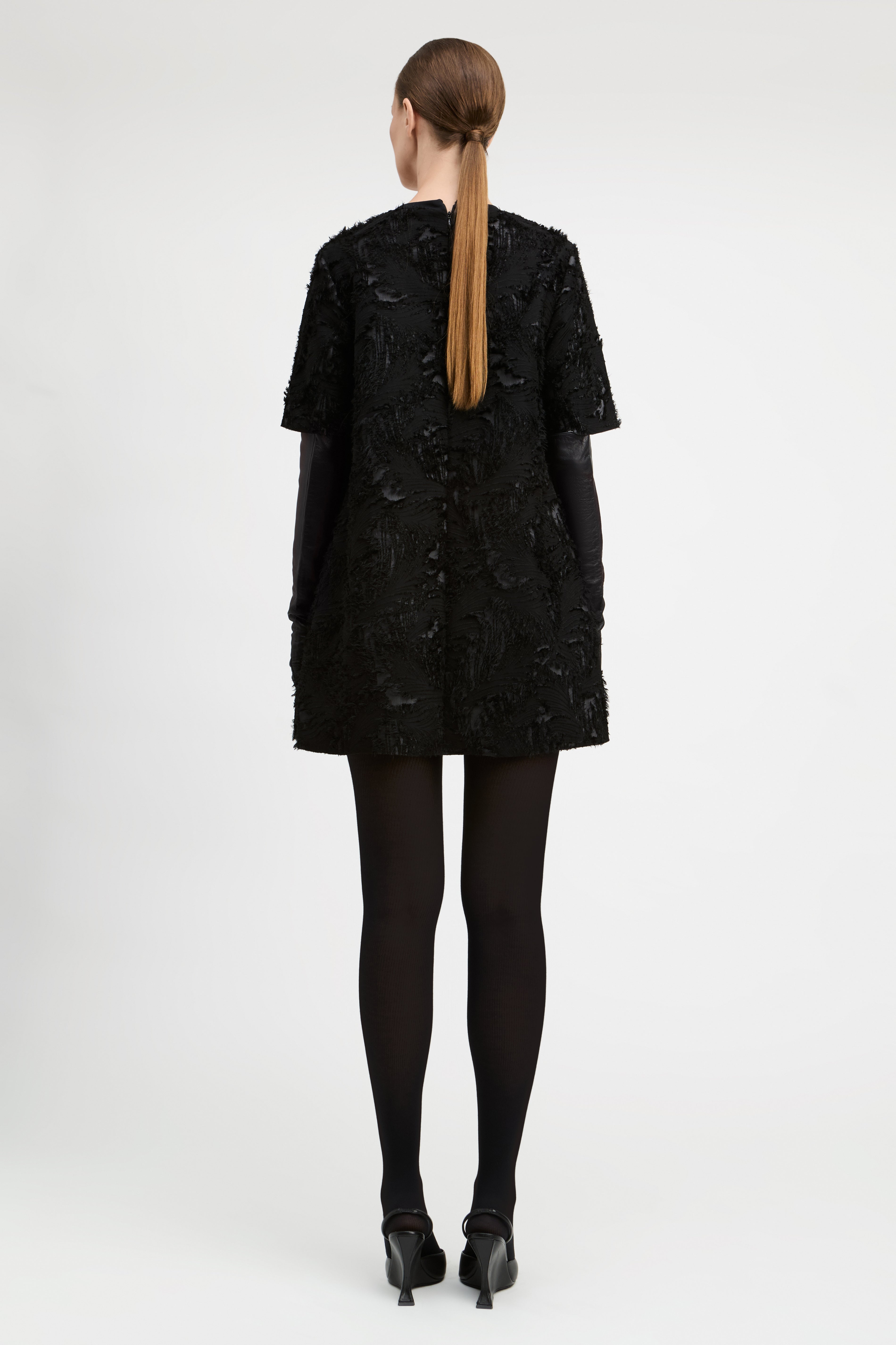 A woman viewed from the back wearing a short black textured dress with black gloves, black tights, and black heels against a plain light gray studio background.