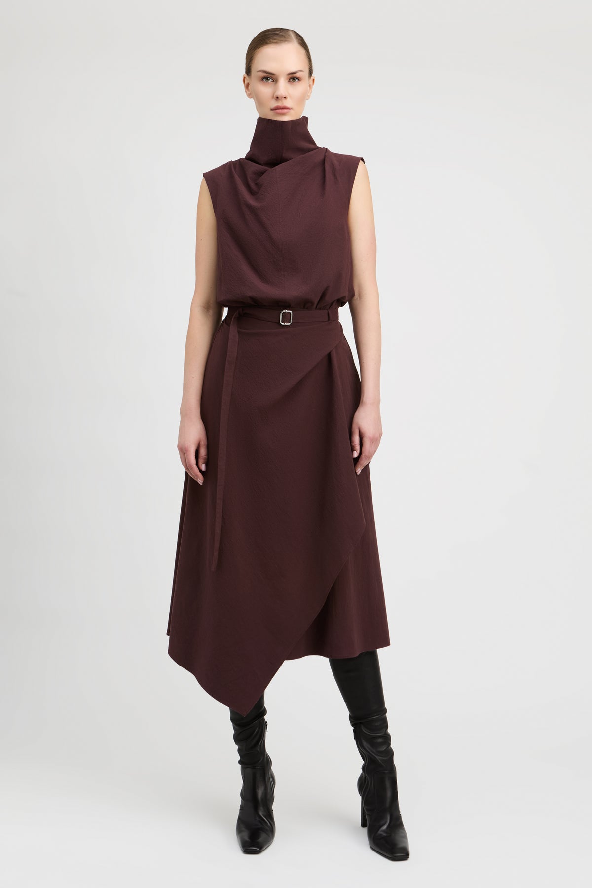 A model stands against a light gray studio background wearing a sleeveless, high-neck, dark brown midi dress with a matching belt and black knee-high boots.