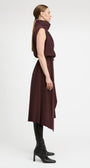 A woman in profile wearing a sleeveless, high-neck, dark plum-brown midi dress with an asymmetrical hem, styled with black knee-high socks and black heeled ankle boots against a light gray studio background.