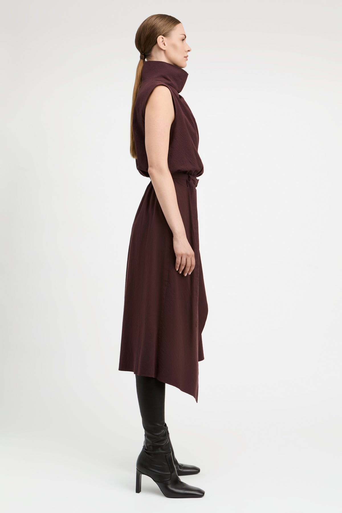 A woman in profile wearing a sleeveless, high-neck, dark plum-brown midi dress with an asymmetrical hem, styled with black knee-high socks and black heeled ankle boots against a light gray studio background.