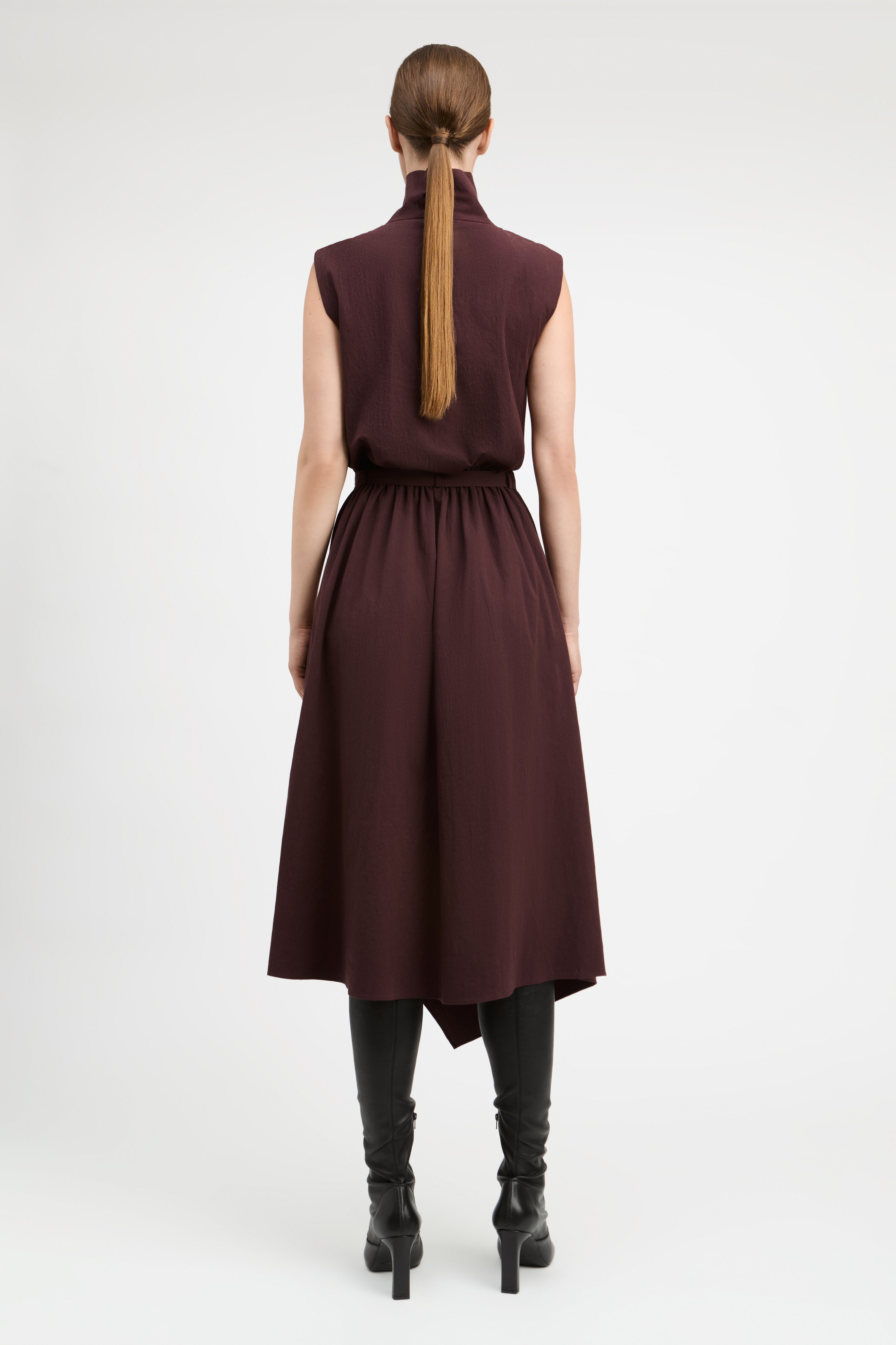A woman viewed from the back wearing a sleeveless high-neck midi dress in deep burgundy, paired with black heeled boots, against a plain light gray studio background.