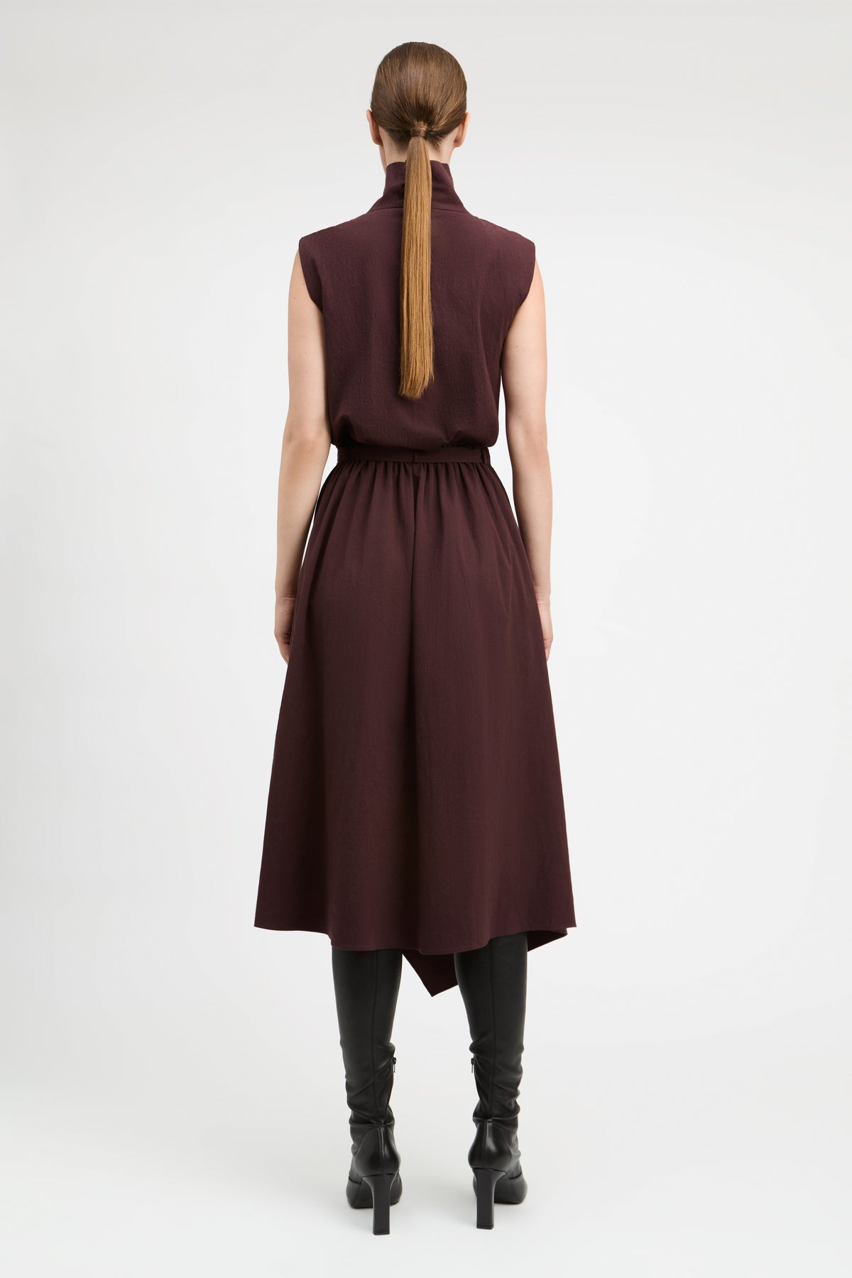 A woman viewed from the back wearing a sleeveless high-neck midi dress in deep burgundy, paired with black heeled boots, against a plain light gray studio background.
