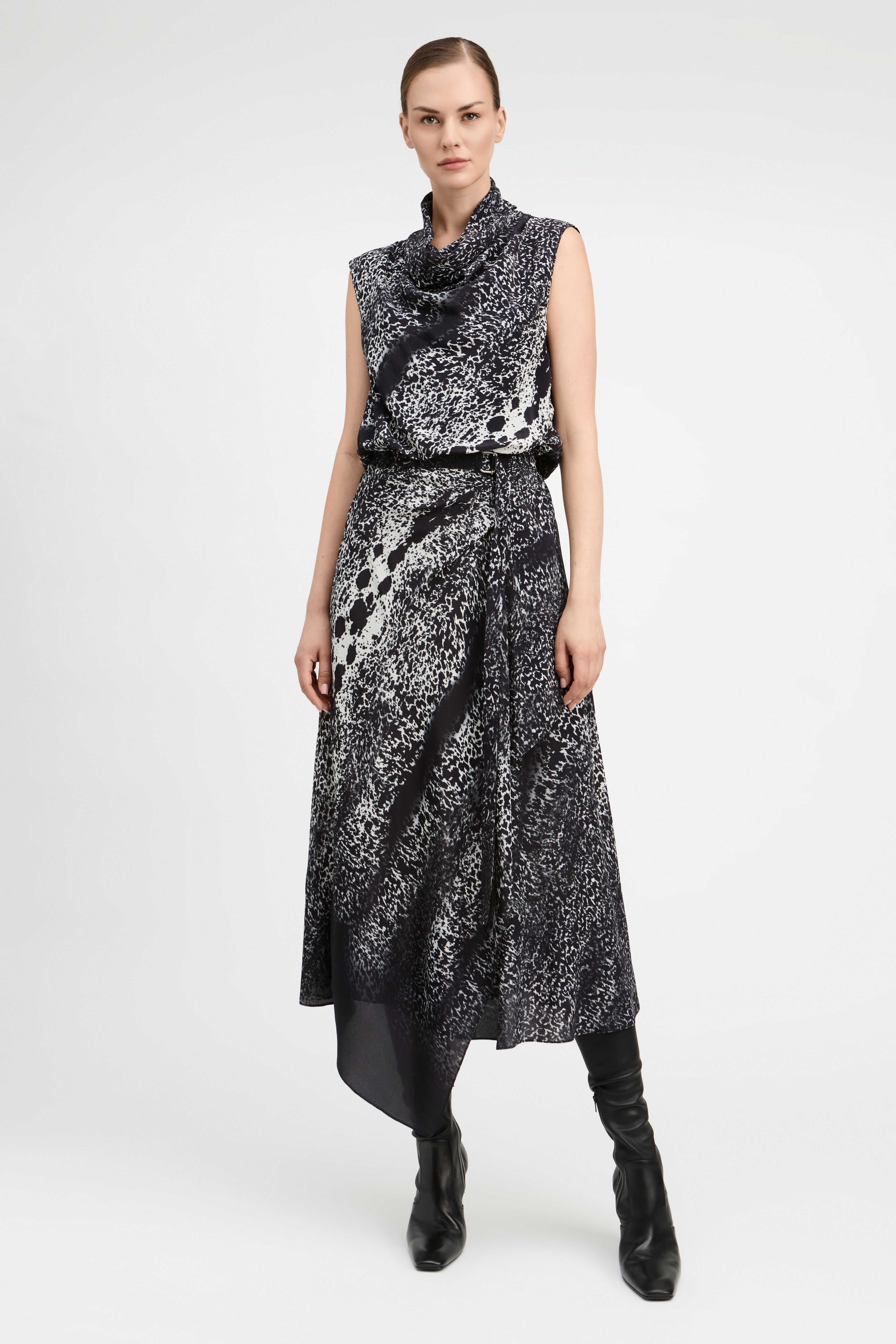 A model wears a sleeveless black and white abstract patterned midi dress with a high cowl neck, a matching black belt, and black knee-high boots, photographed against a light gray studio background.