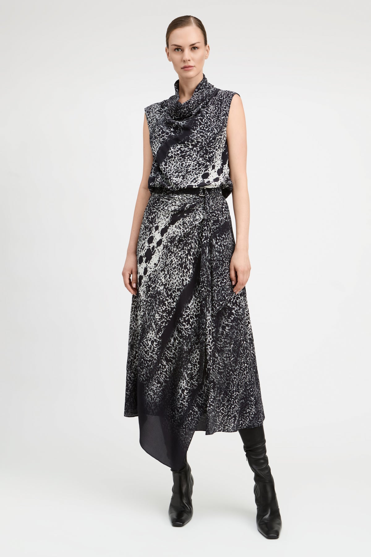 A model wears a sleeveless black and white abstract patterned midi dress with a high cowl neck, a matching black belt, and black knee-high boots, photographed against a light gray studio background.