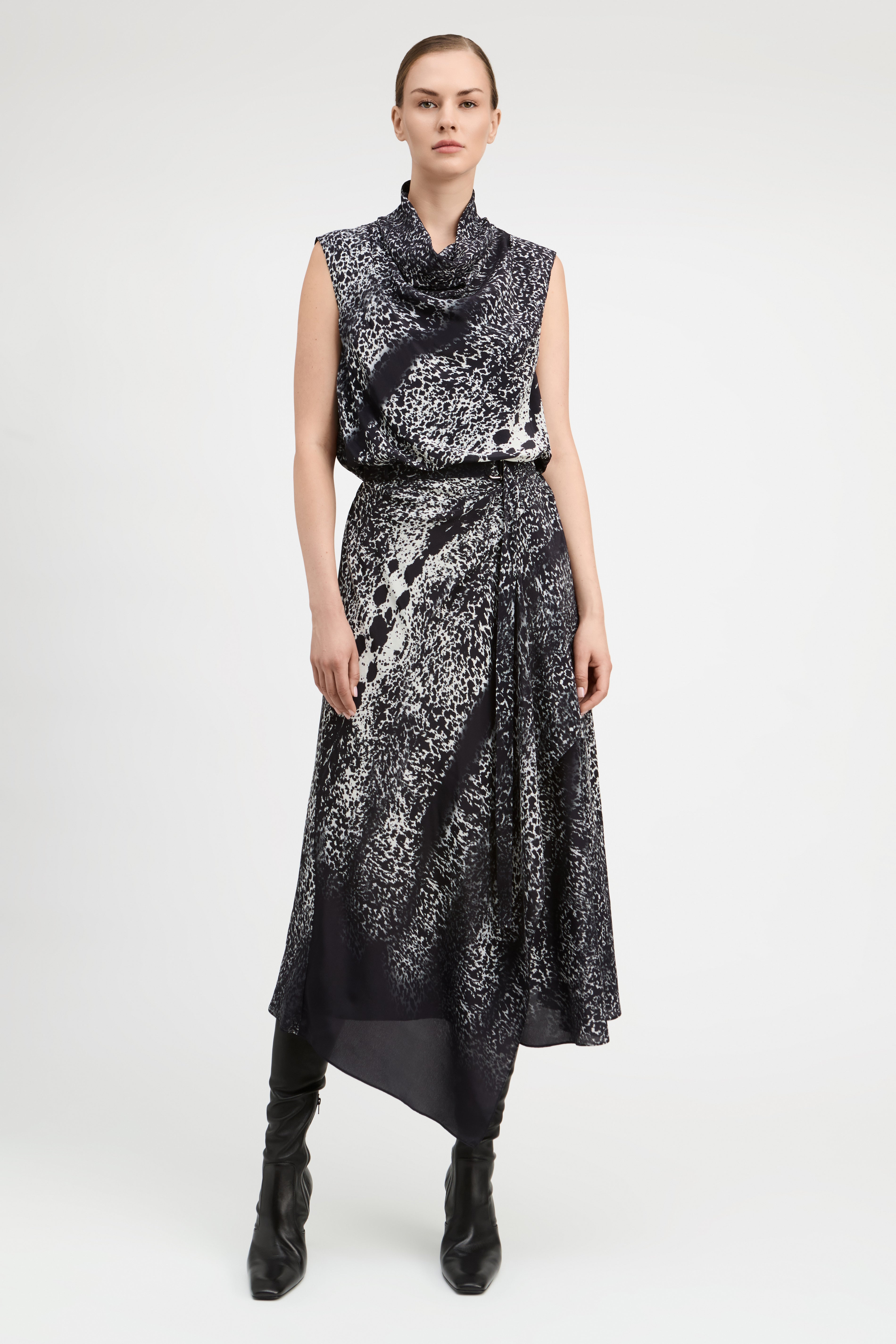A model stands against a light gray studio background wearing a sleeveless, high-neck midi dress with an abstract black and white pattern and black pointed boots.