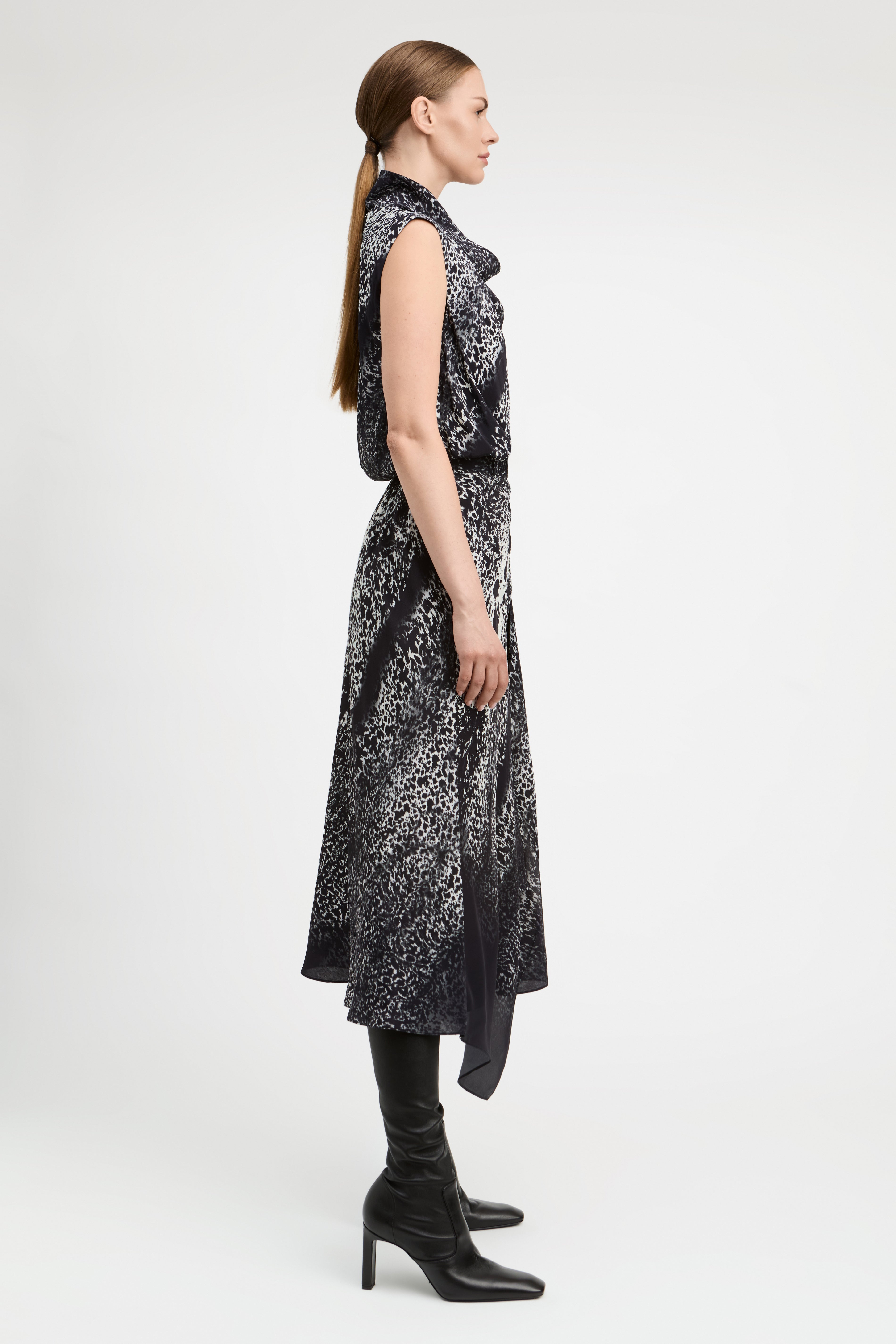 A woman in profile wearing a sleeveless black midi dress with a white abstract pattern, paired with black knee-high boots, against a plain light gray studio background.