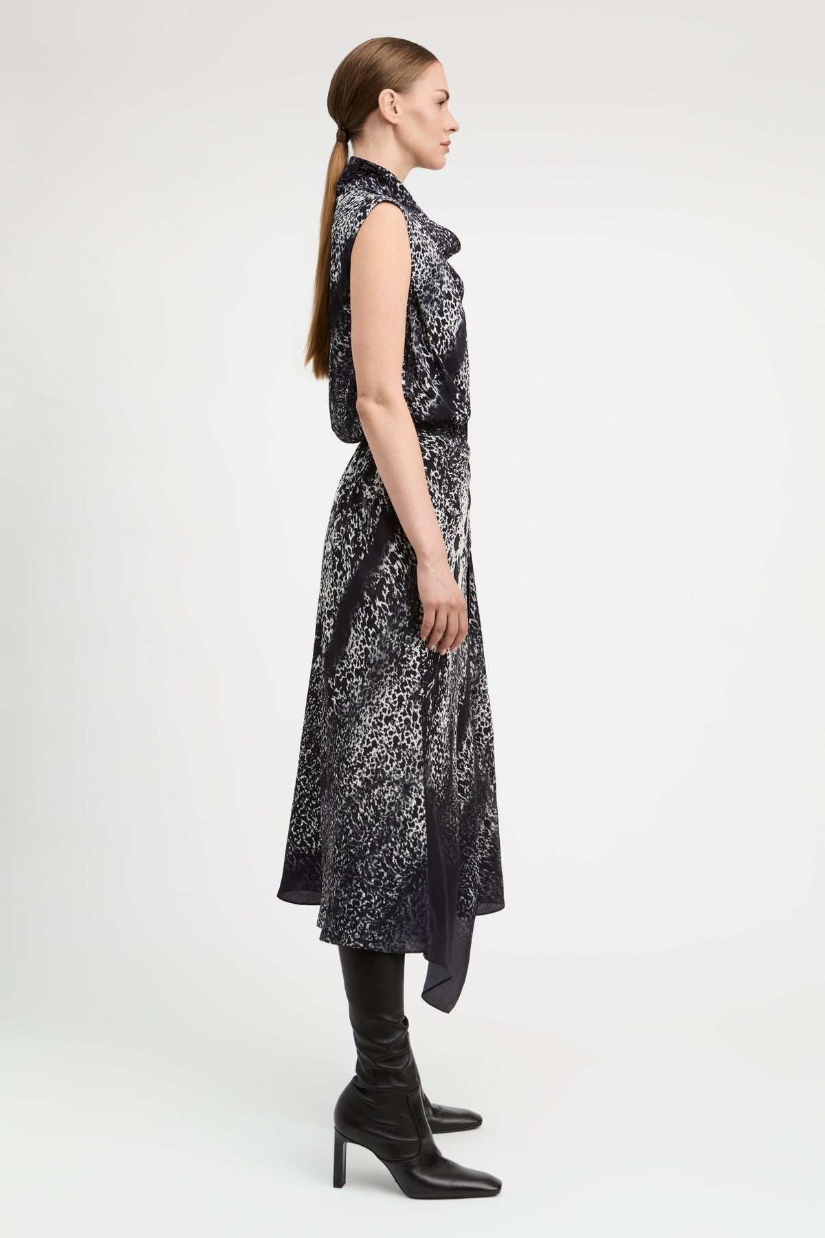 A woman in profile wearing a sleeveless black midi dress with a white abstract pattern, paired with black knee-high boots, against a plain light gray studio background.