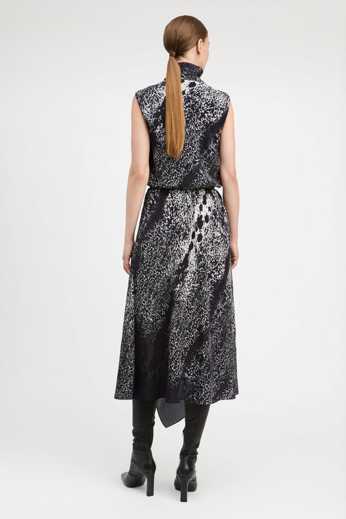 A woman viewed from the back wearing a sleeveless high-neck midi dress with a black and white abstract pattern, paired with black knee-high boots, against a light gray studio background.