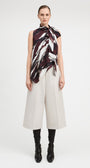 A model stands against a light gray studio background wearing a sleeveless high-neck scarf top with a dark abstract print, off-white cropped wide-leg pants, and black knee-high boots.