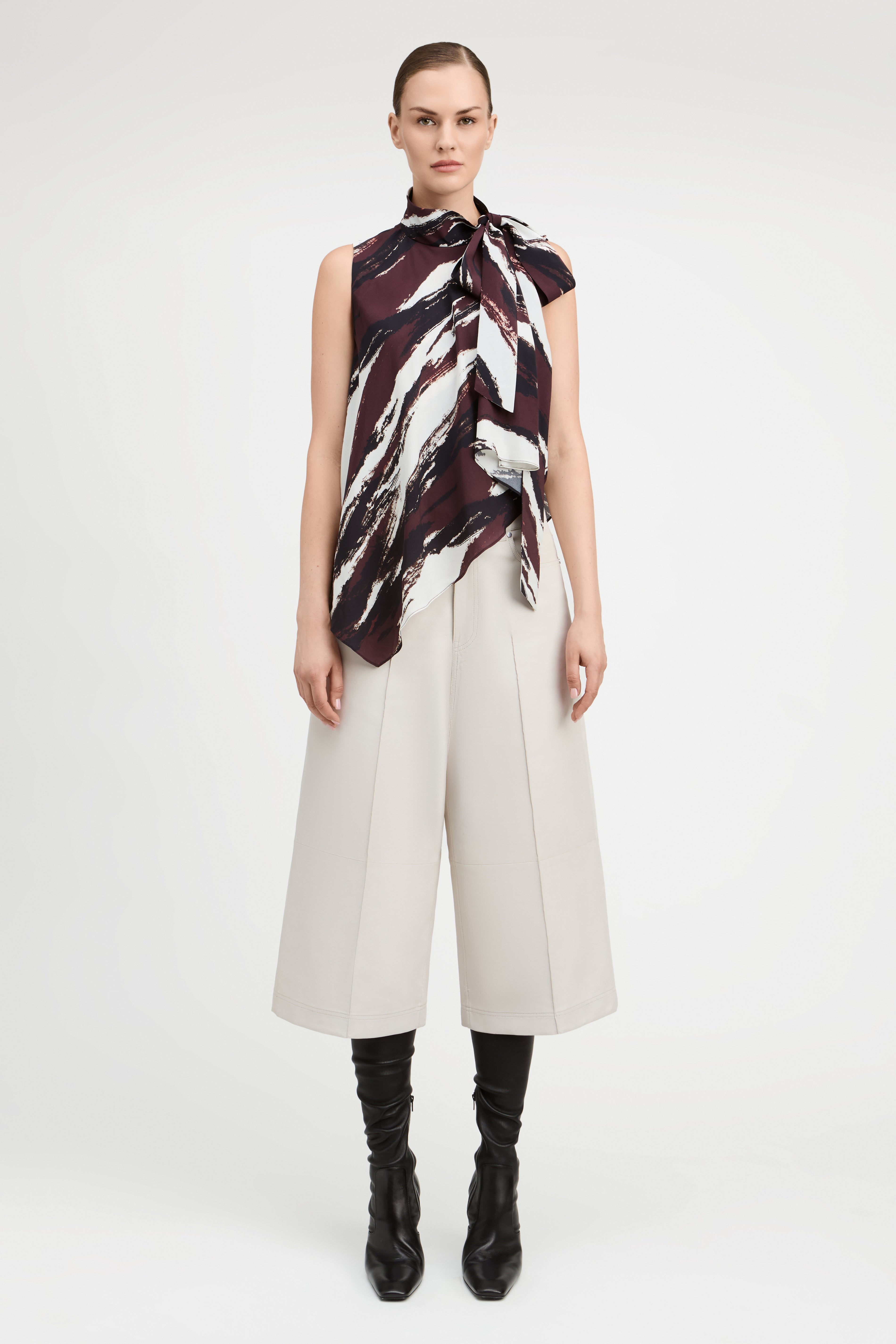 A model stands against a light gray studio background wearing a sleeveless high-neck scarf top with a dark abstract print, off-white cropped wide-leg pants, and black knee-high boots.
