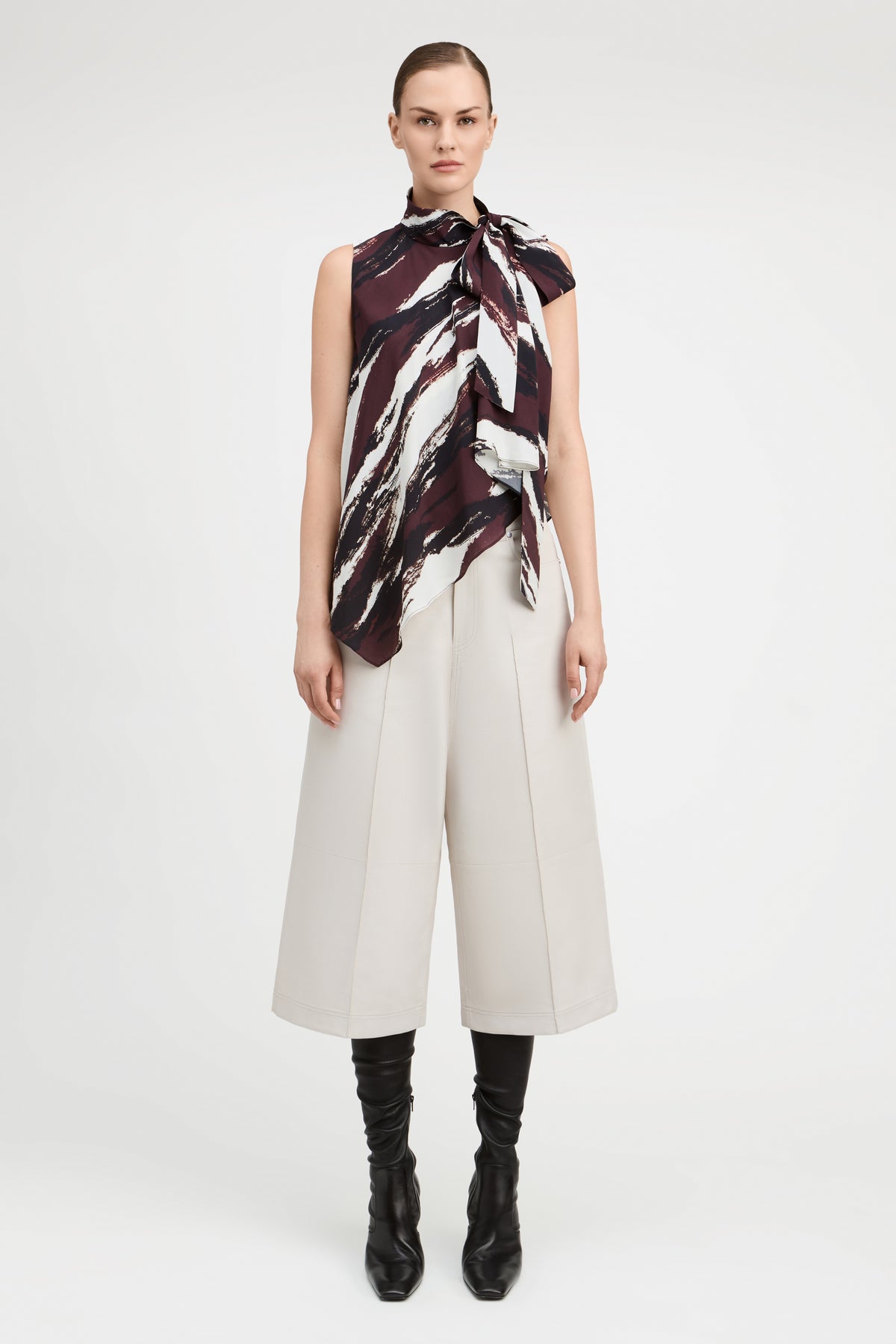 A model stands against a light gray studio background wearing a sleeveless high-neck scarf top with a dark abstract print, off-white cropped wide-leg pants, and black knee-high boots.