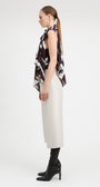 A woman stands in profile wearing a sleeveless top with a dark abstract print, a long cream skirt, black knee-high socks, and black heeled boots against a plain light gray studio background.