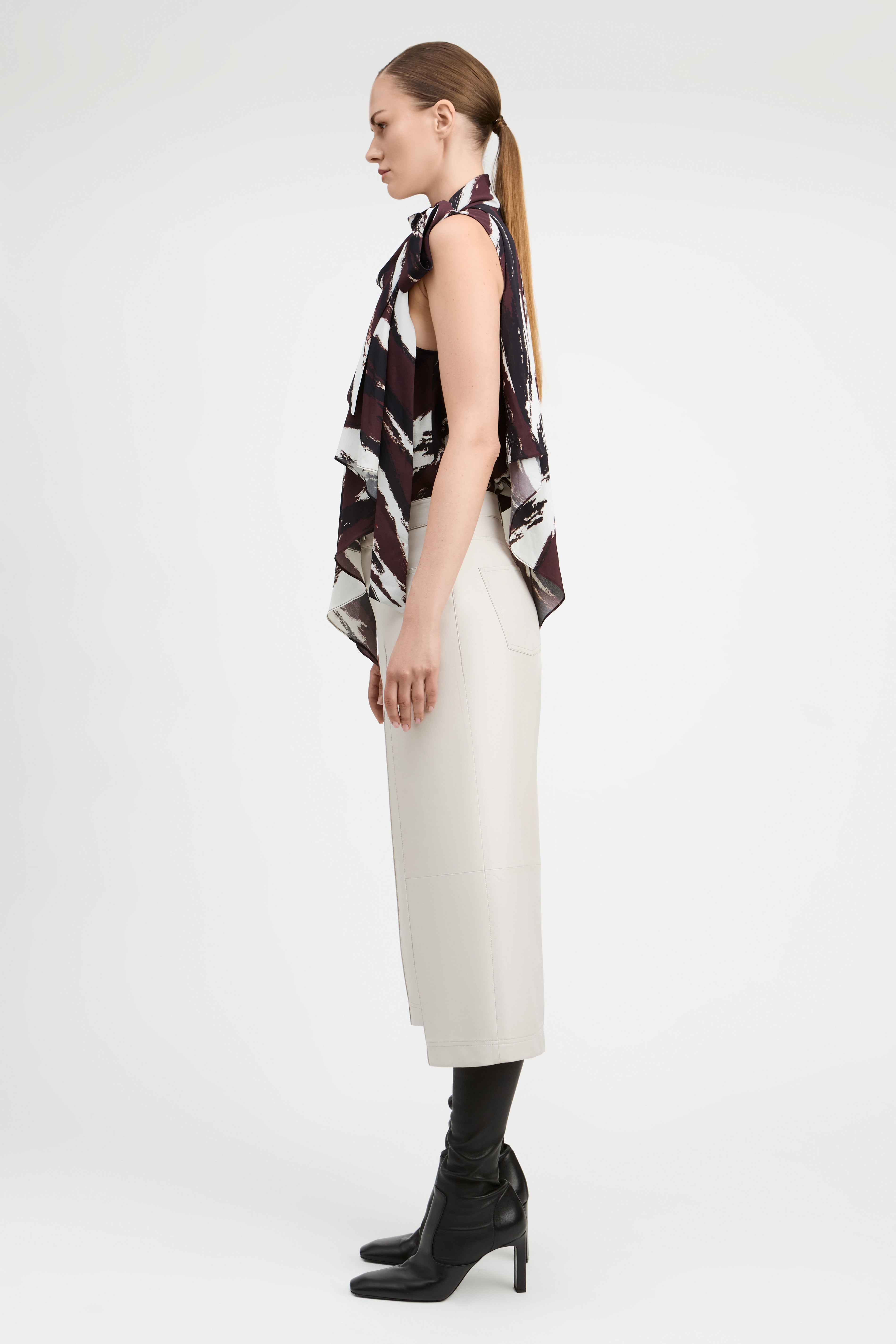 A woman stands in profile wearing a sleeveless top with a dark abstract print, a long cream skirt, black knee-high socks, and black heeled boots against a plain light gray studio background.