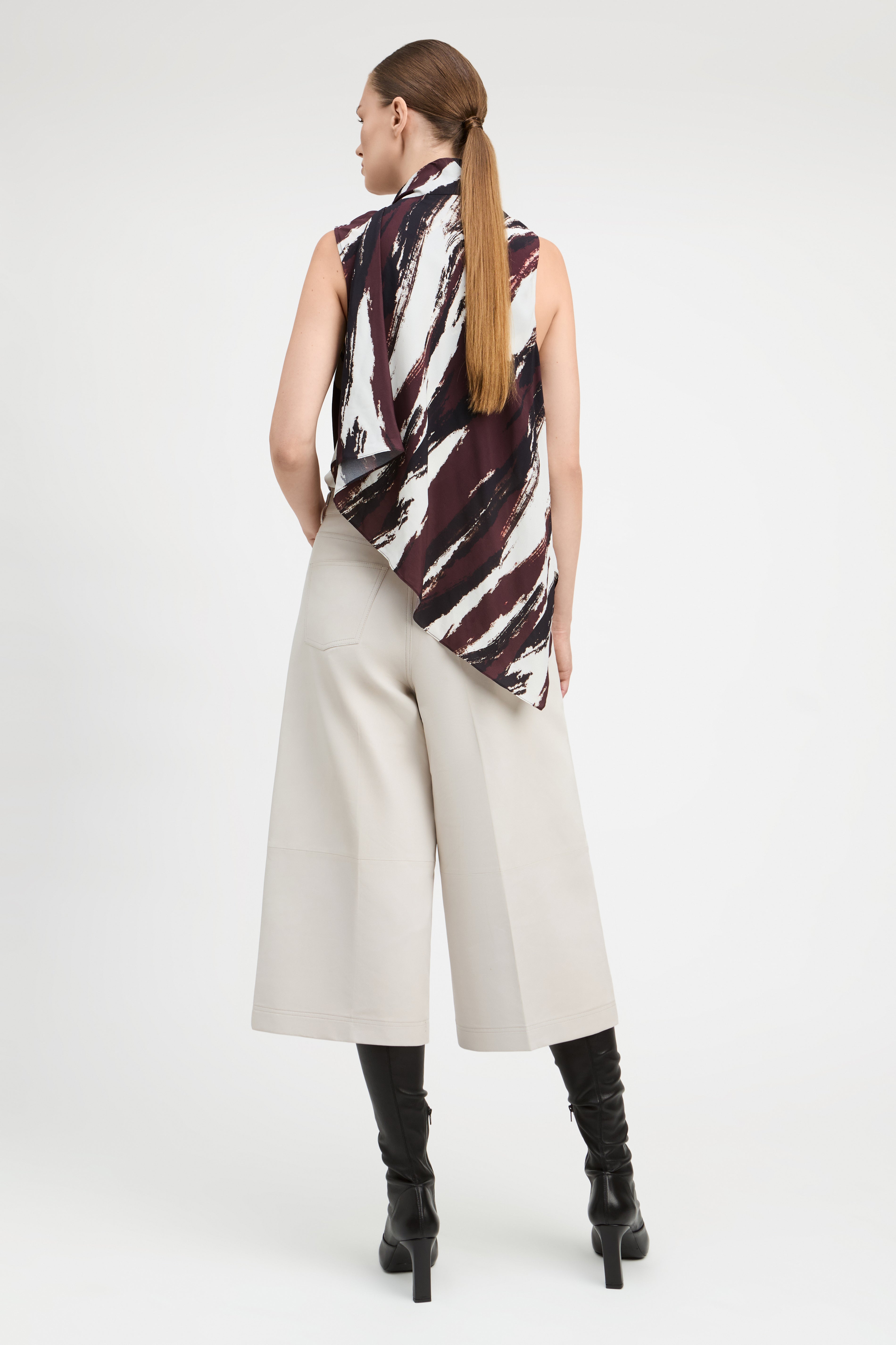 A woman is shown from the back wearing a sleeveless top with a black, white, and dark red abstract brushstroke pattern, paired with light beige wide-leg cropped pants and black knee-high boots, against a plain white studio background.
