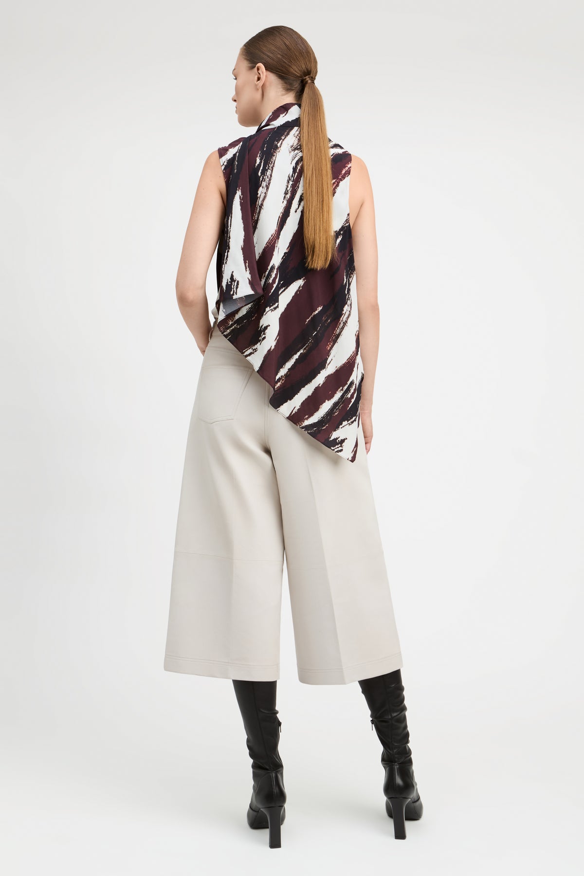 A woman is shown from the back wearing a sleeveless top with a black, white, and dark red abstract brushstroke pattern, paired with light beige wide-leg cropped pants and black knee-high boots, against a plain white studio background.