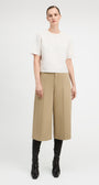 A model stands against a light gray studio background wearing a textured off-white short-sleeve top, beige wide-leg cropped trousers, and black knee-high boots.