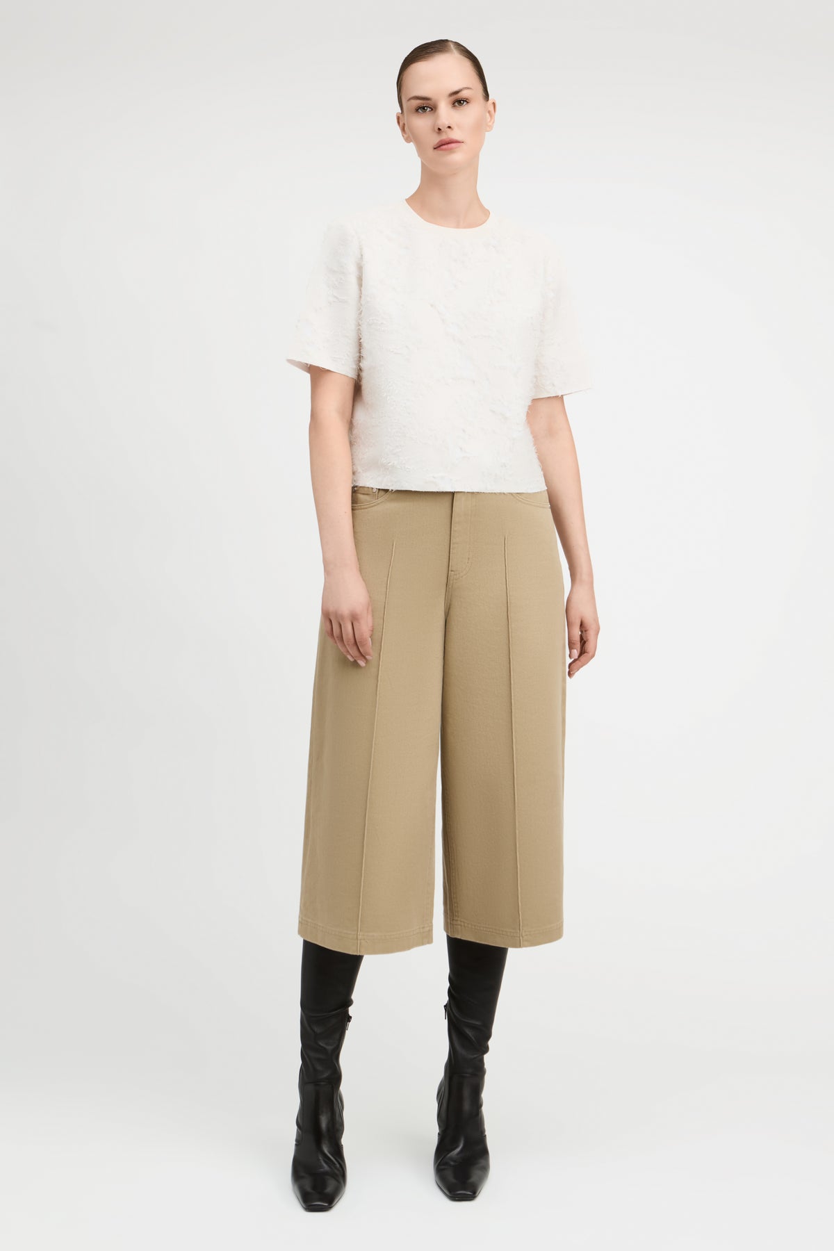 A model stands against a light gray studio background wearing a textured off-white short-sleeve top, beige wide-leg cropped trousers, and black knee-high boots.