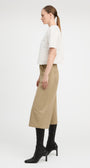A woman shown in profile wearing a white textured short-sleeve top, beige cropped trousers, and black heeled ankle boots against a plain light gray studio background.
