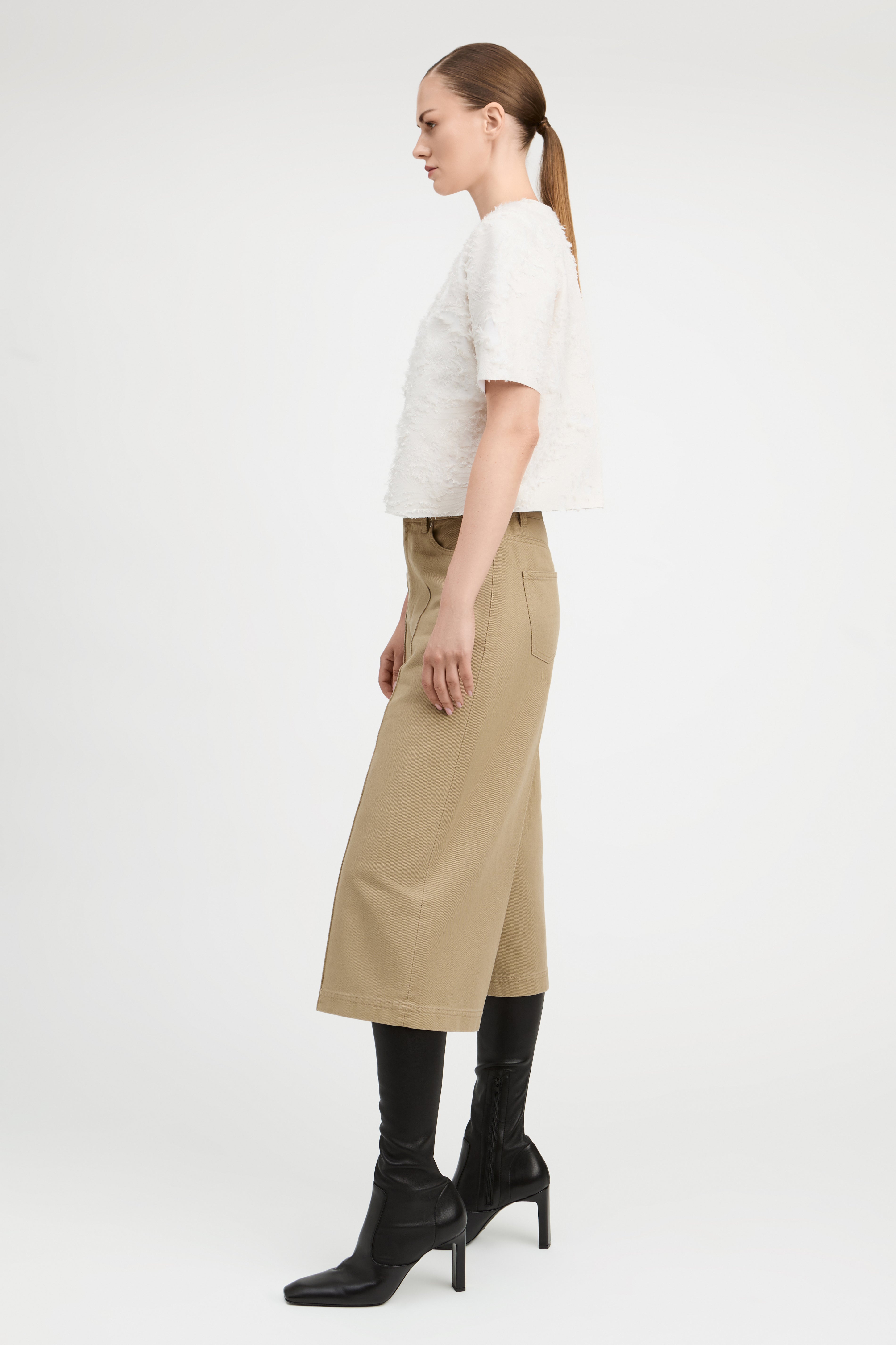 A woman shown in profile wearing a white textured short-sleeve top, beige cropped trousers, and black heeled ankle boots against a plain light gray studio background.