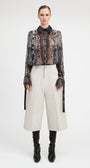 A model stands against a light gray studio background wearing a sheer black and brown patterned long-sleeve blouse with a high ruffled collar, paired with off-white wide-leg cropped trousers, black knee-high socks, and black ankle boots.