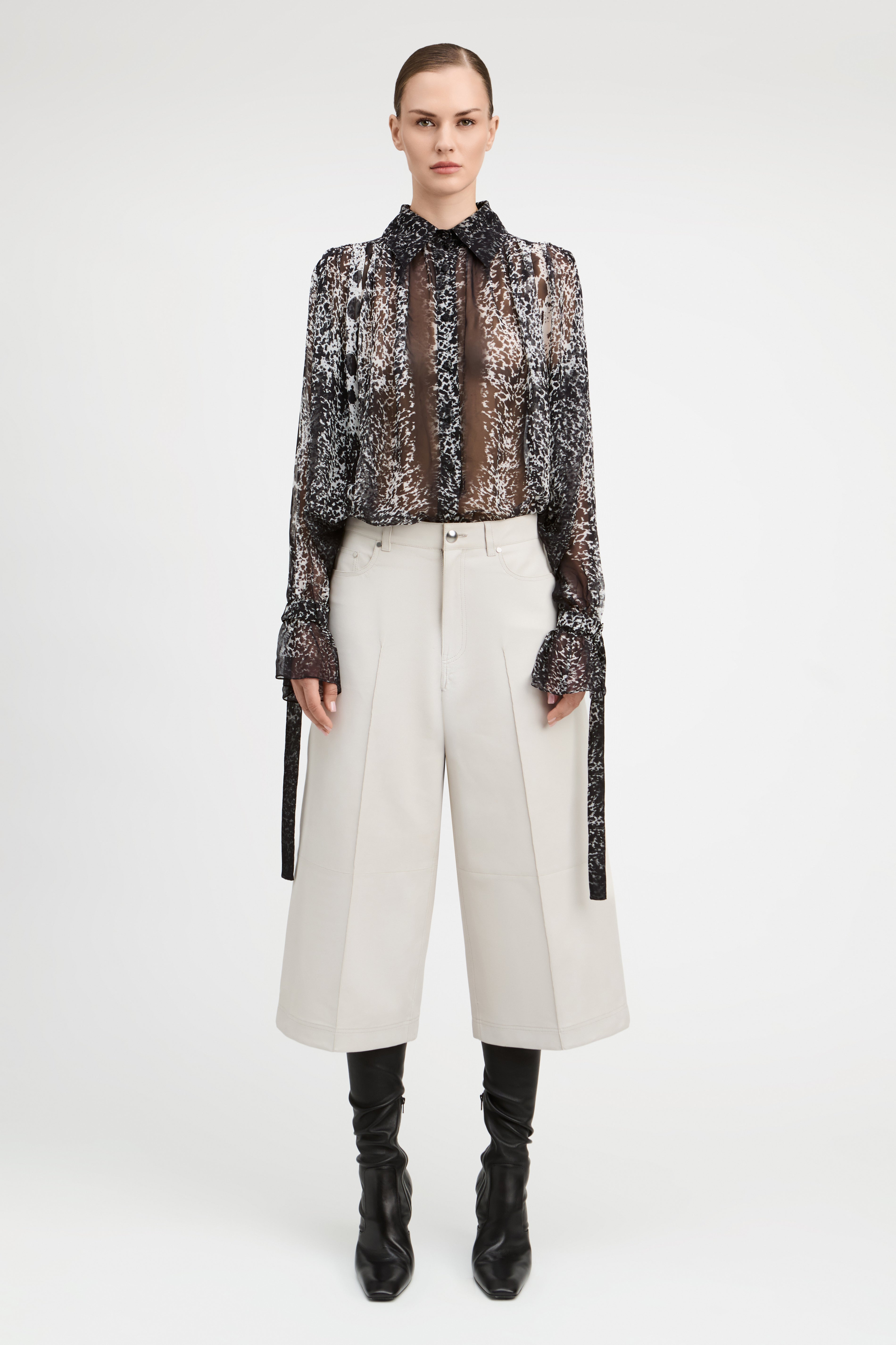 A model stands against a light gray studio background wearing a sheer black and brown patterned long-sleeve blouse with a high ruffled collar, paired with off-white wide-leg cropped trousers, black knee-high socks, and black ankle boots.