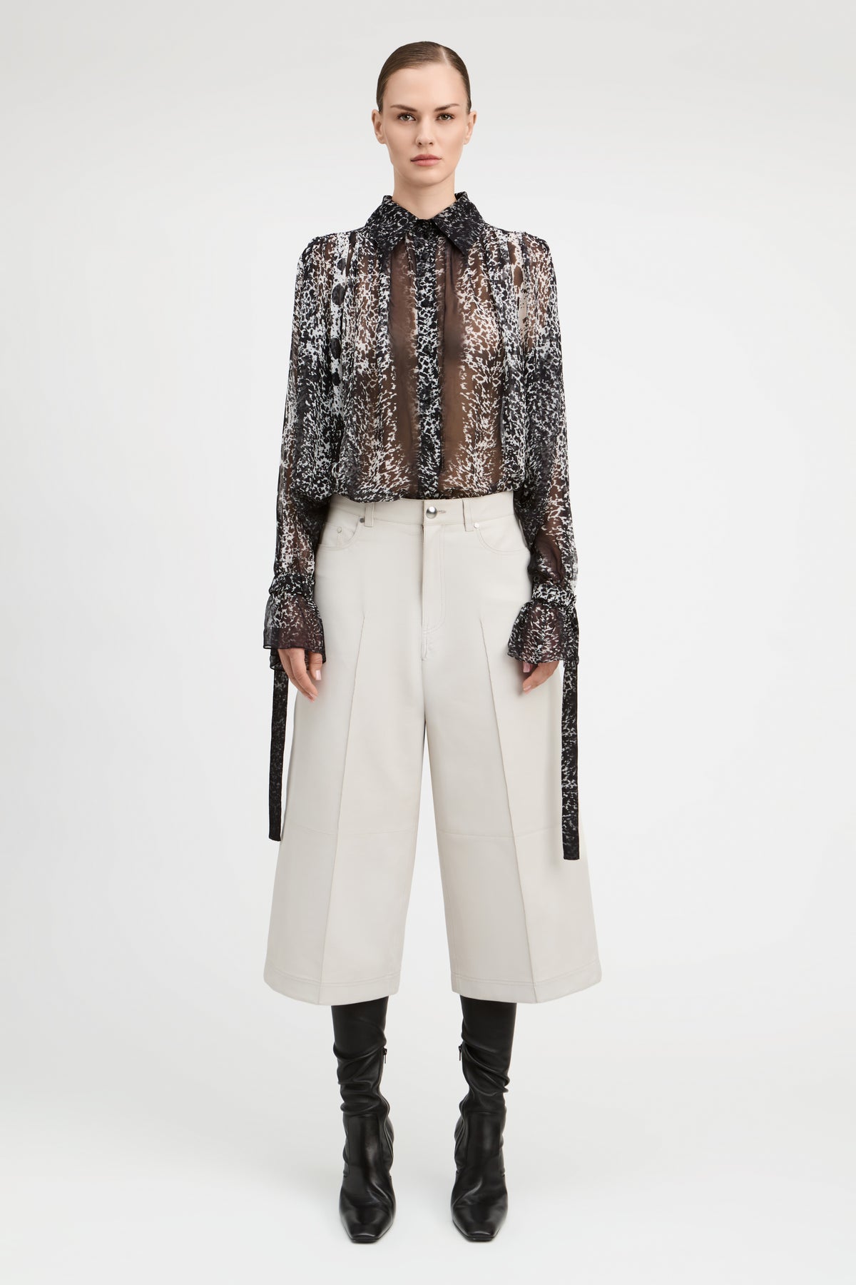 A model stands against a light gray studio background wearing a sheer black and brown patterned long-sleeve blouse with a high ruffled collar, paired with off-white wide-leg cropped trousers, black knee-high socks, and black ankle boots.
