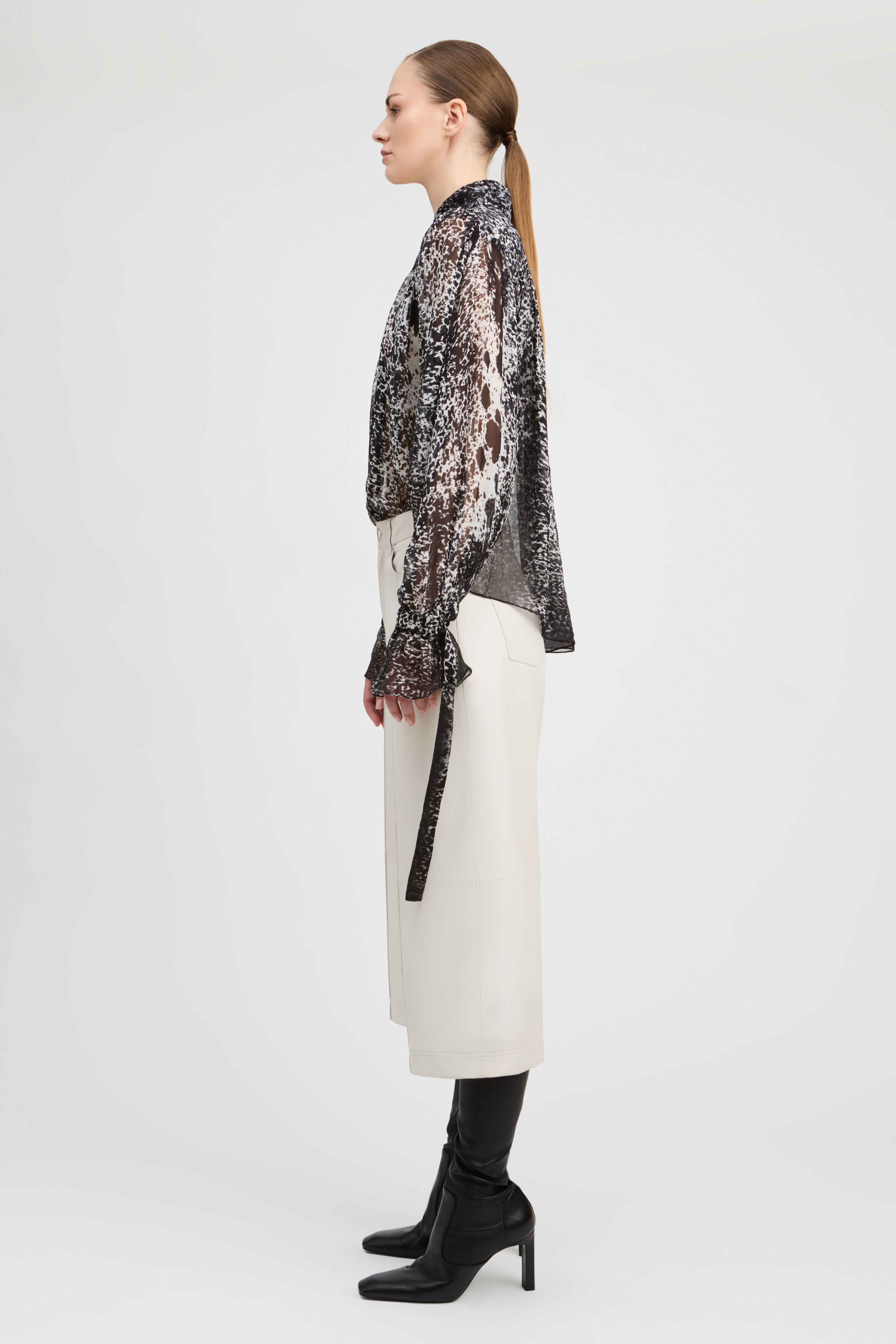 A model shown in profile wearing a sheer black and white patterned long-sleeve top, a white midi skirt, and black heeled boots, photographed against a plain light gray studio background.