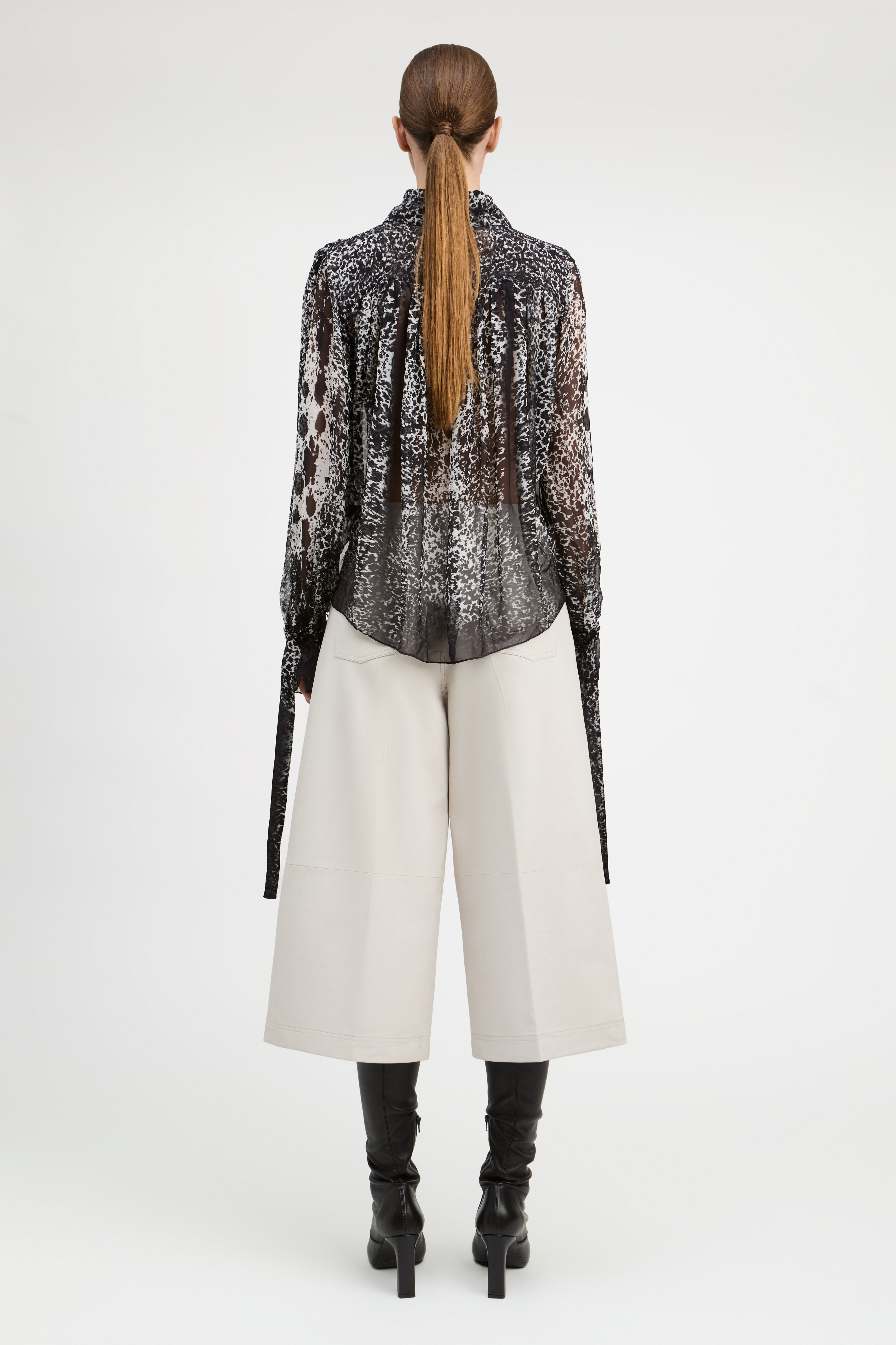 A model shown from the back wearing a sheer black and white patterned long-sleeve blouse, off-white wide-leg cropped pants, and black heeled boots against a light gray studio background.