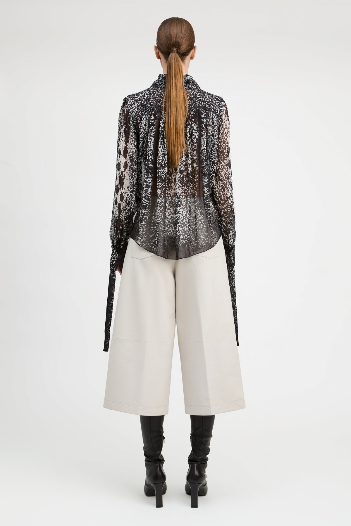 A model shown from the back wearing a sheer black and white patterned long-sleeve blouse, off-white wide-leg cropped pants, and black heeled boots against a light gray studio background.