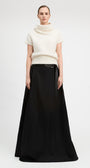 A model stands against a light gray studio background wearing a cream short-sleeve mock-neck knit top and a long black skirt with a black belt.