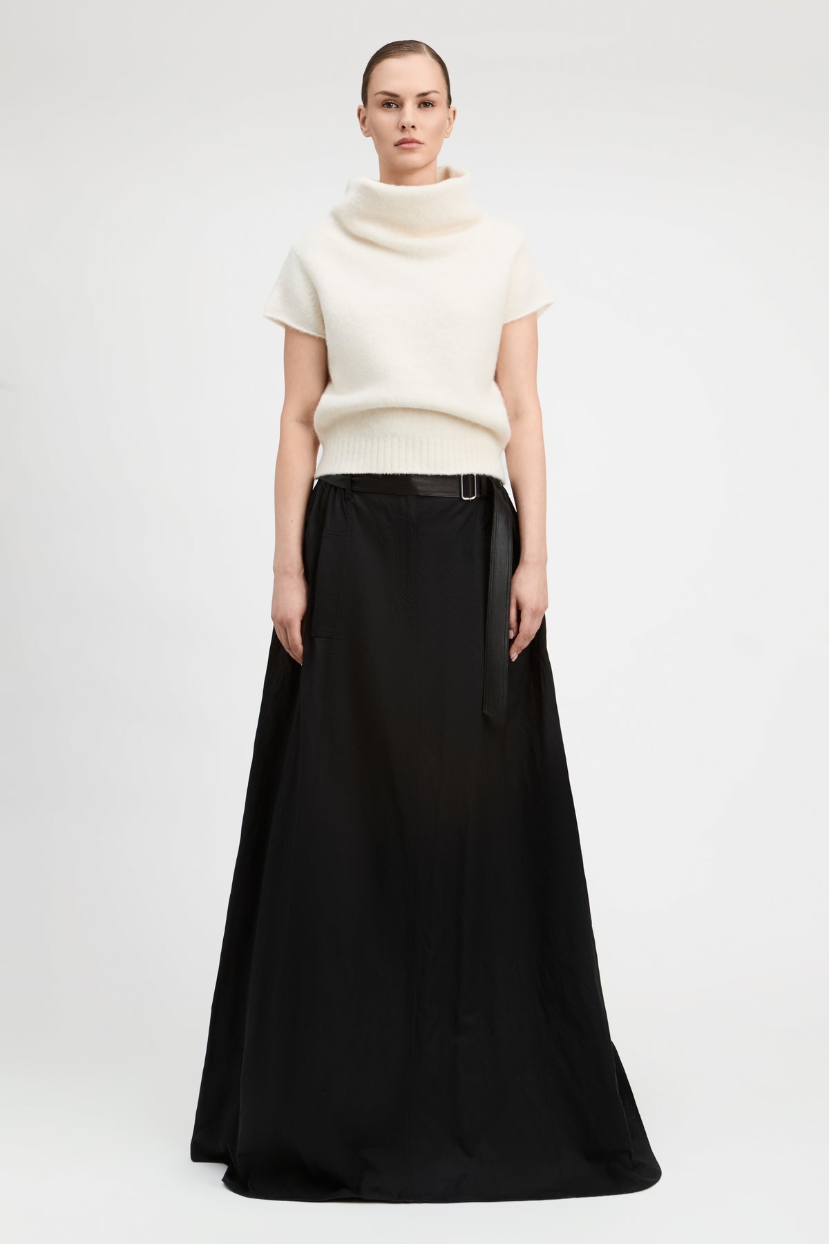 A model stands against a light gray studio background wearing a cream short-sleeve mock-neck knit top and a long black skirt with a black belt.