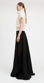A woman in profile wearing a cream short-sleeve mock-neck knit top and a long black skirt, standing against a plain light gray studio background.