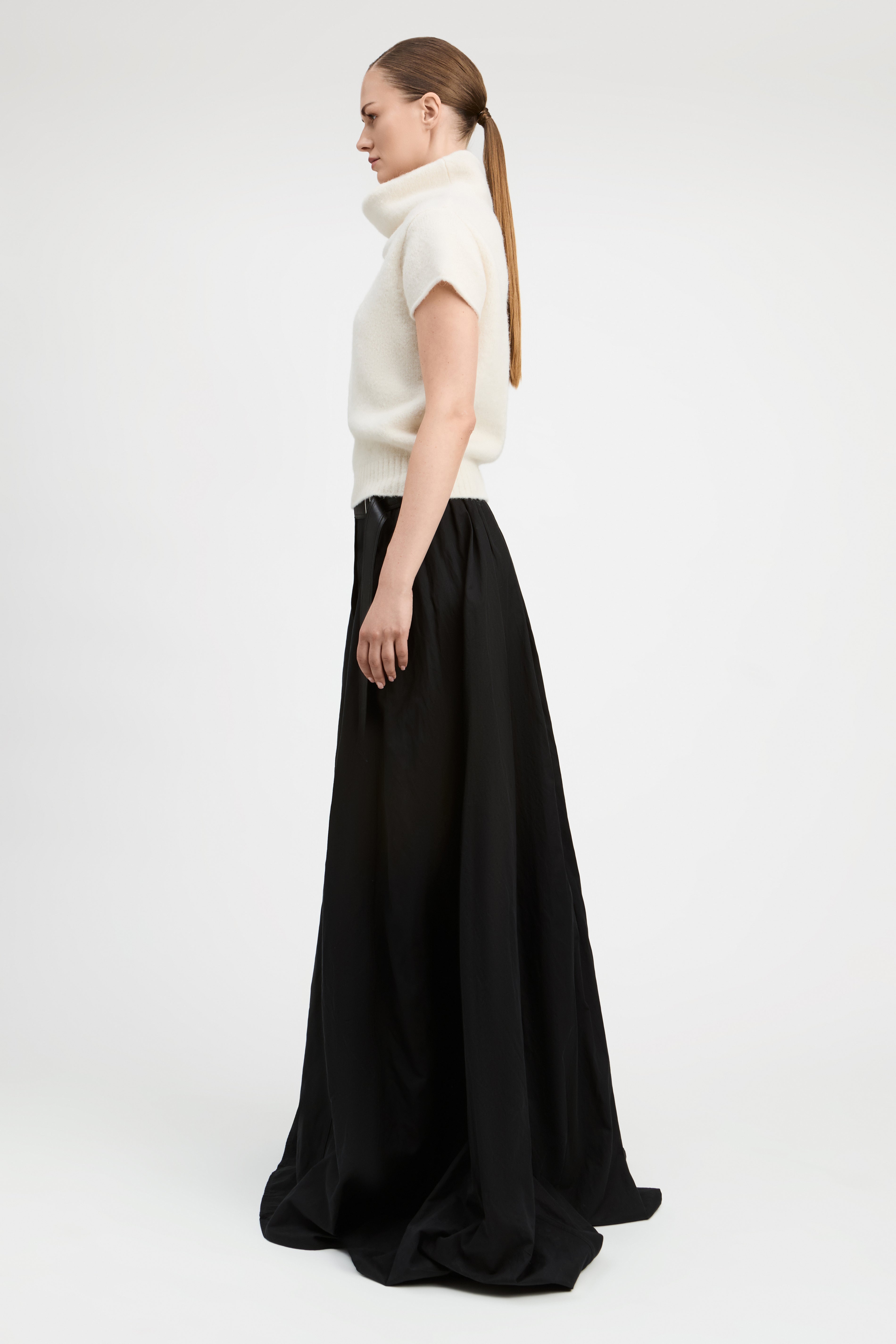 A woman in profile wearing a cream short-sleeve mock-neck knit top and a long black skirt, standing against a plain light gray studio background.